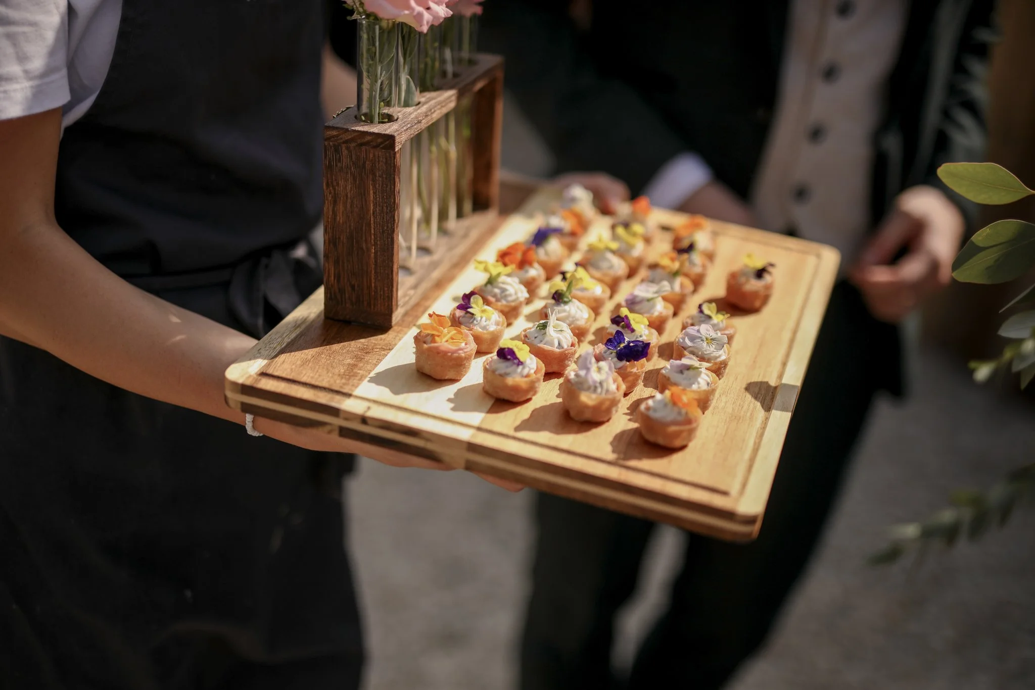 Person holding a wooden tray with colorful appetizer bites topped with edible flowers, with floral arrangements in vases on a wooden stand in the background.