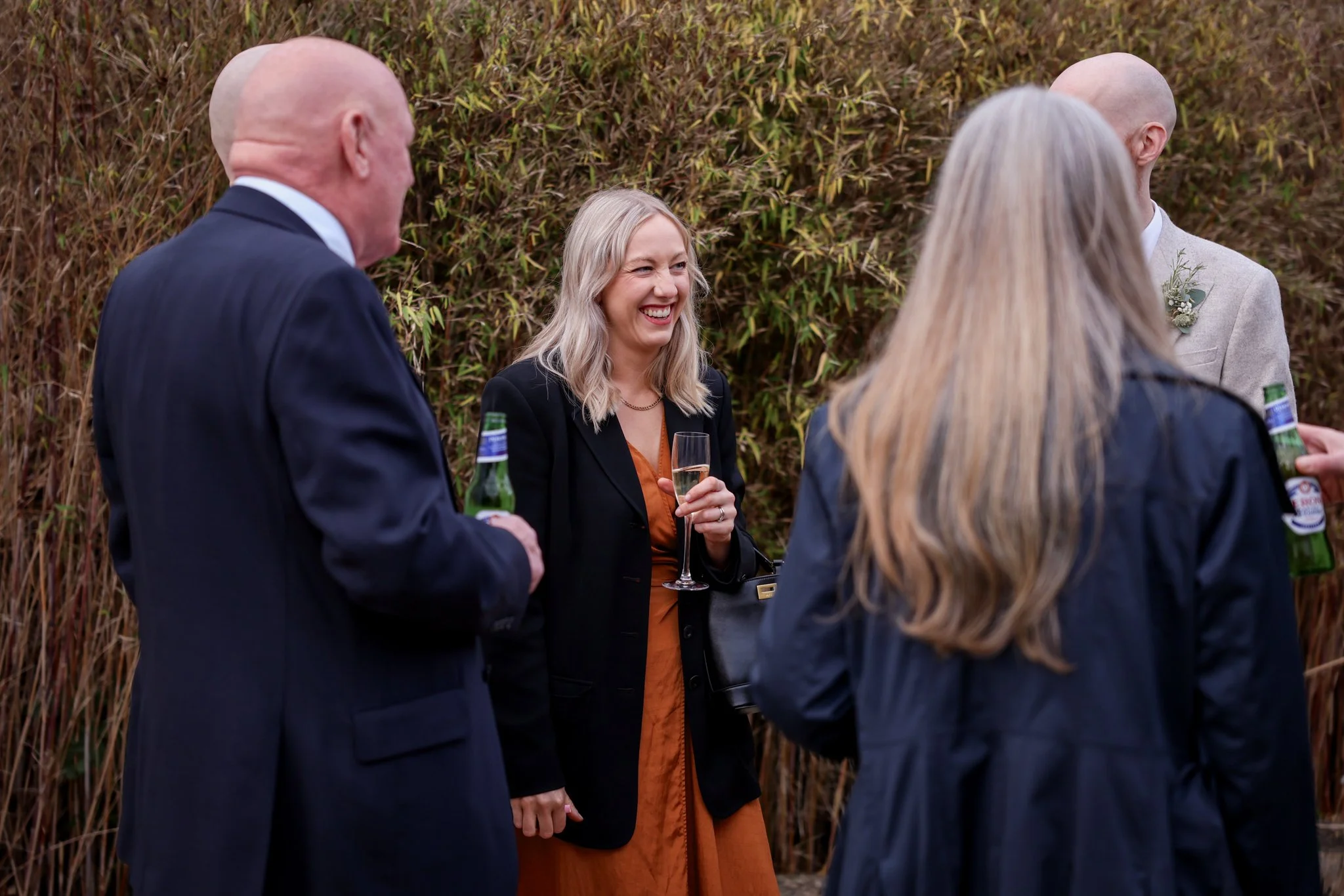 Group of people at an outdoor social gathering, smiling and chatting, with a woman holding a glass of champagne in front of a grassy background.