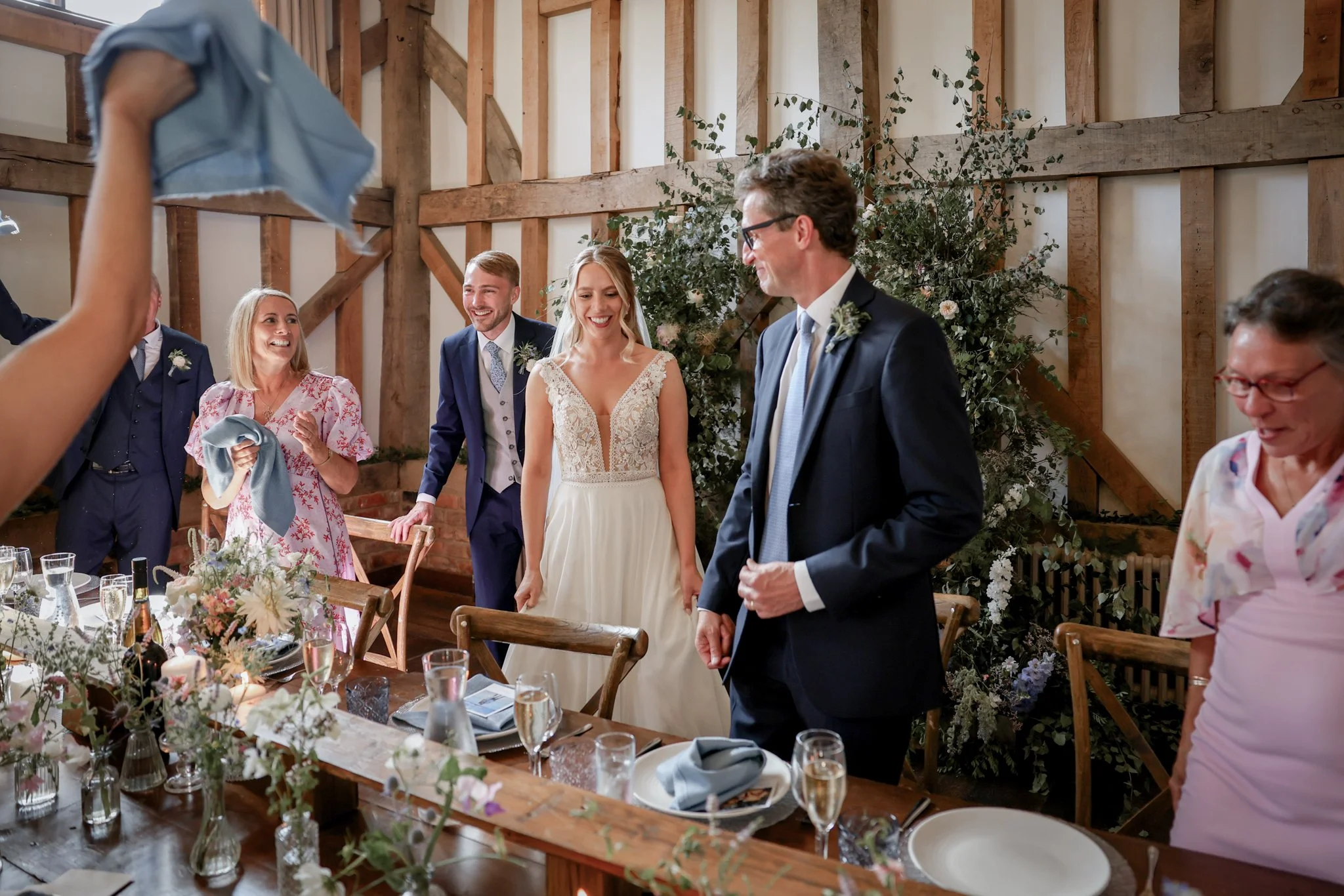 Bride and groom standing at wedding reception, surrounded by friends and family, with a rustic wood-paneled background and floral decorations.