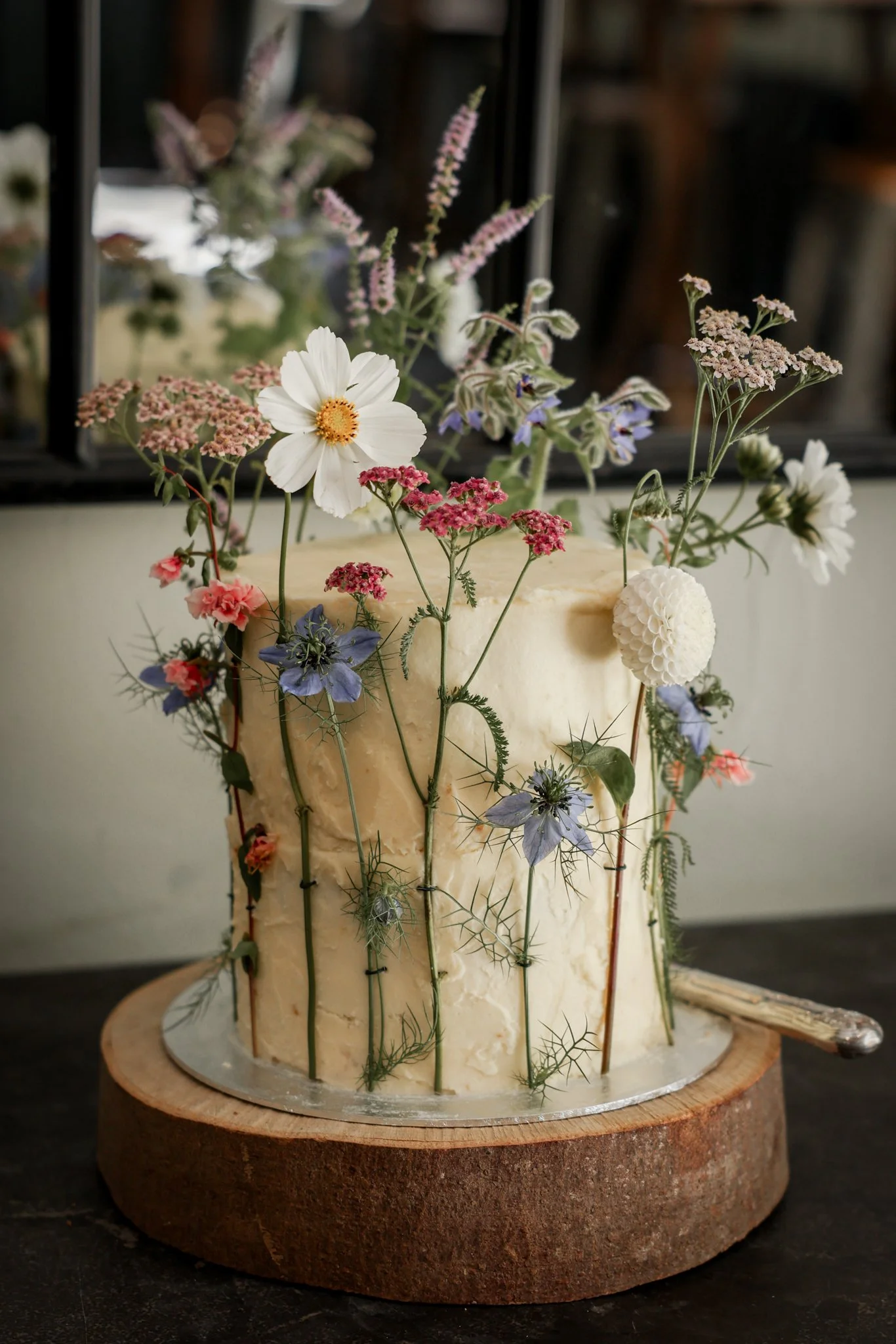 A cheese wheel decorated with various fresh flowers, including white, pink, purple, and blue blooms, mounted on a wooden base.