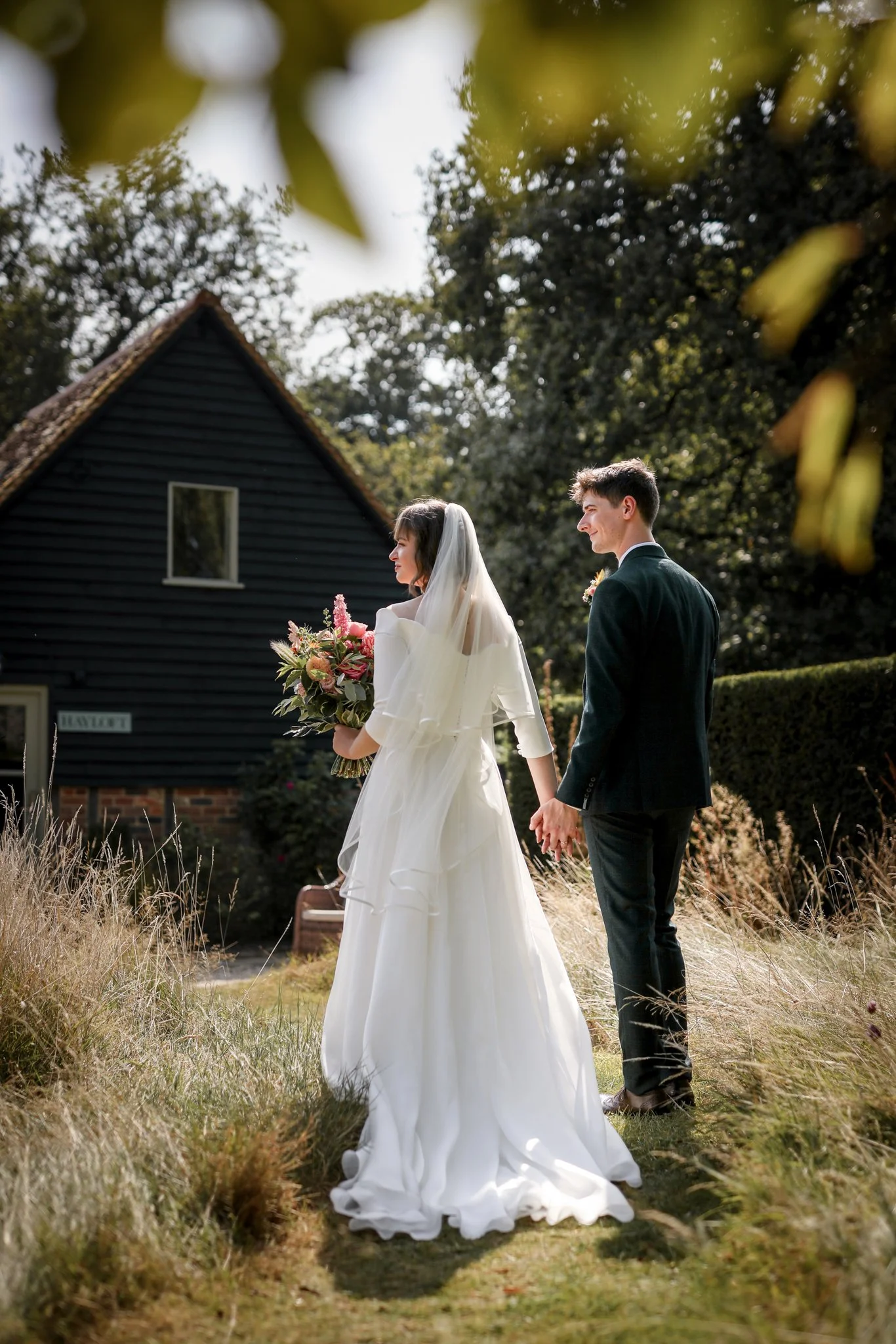 A bride and groom holding hands outdoors, the bride in a white wedding dress holding a bouquet, and the groom in a dark suit, walking through a grassy area near a black house with a steep roof.