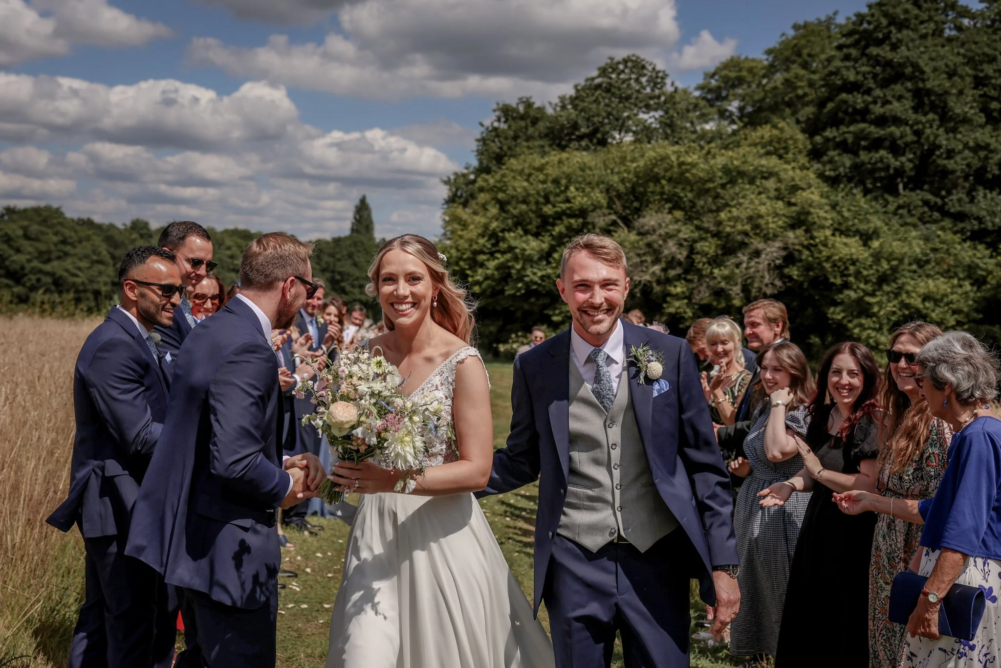 A wedding ceremony outdoors with a bride and groom holding hands, surrounded by guests during the daytime.
