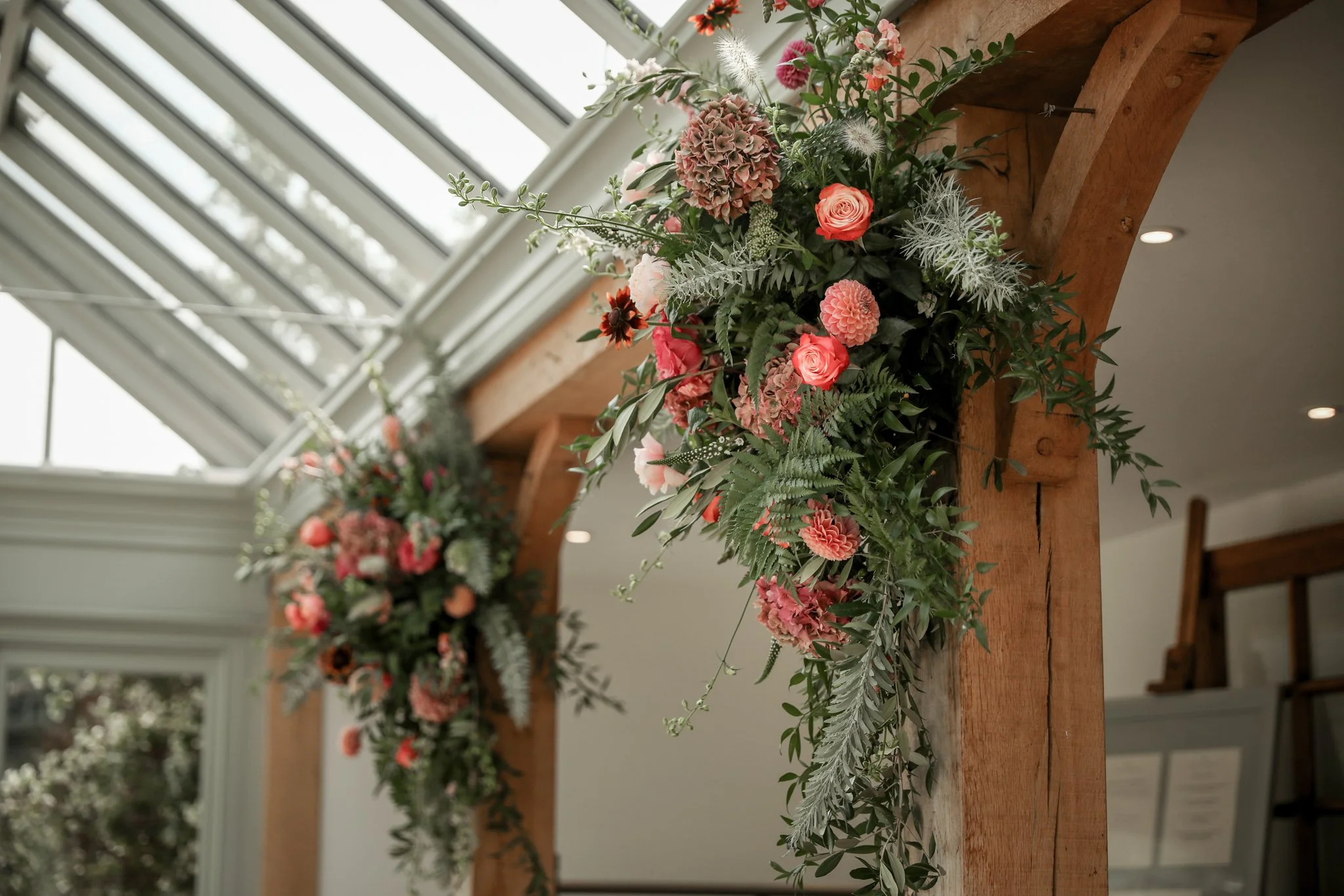A floral arrangement with pink and peach roses, hydrangeas, and assorted greenery on a wooden table in a room with a sunlit window and ceiling lights.