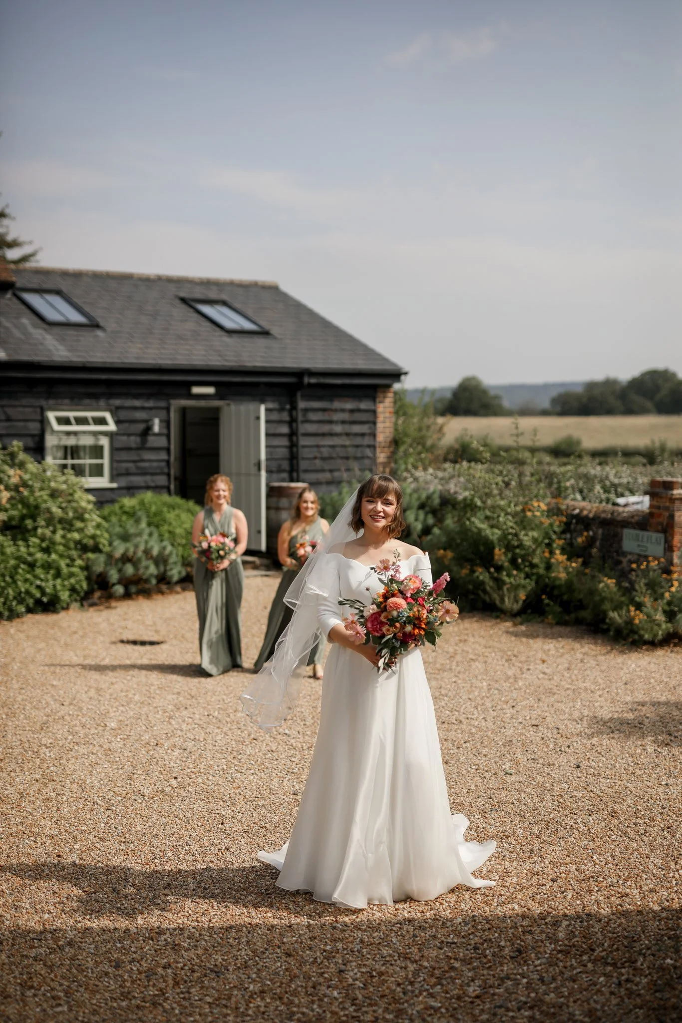 A bride in a white wedding dress holding a bouquet of flowers, smiling outdoors with two bridesmaids in grey dresses holding bouquets in the background, beside a black barn in a rural setting.