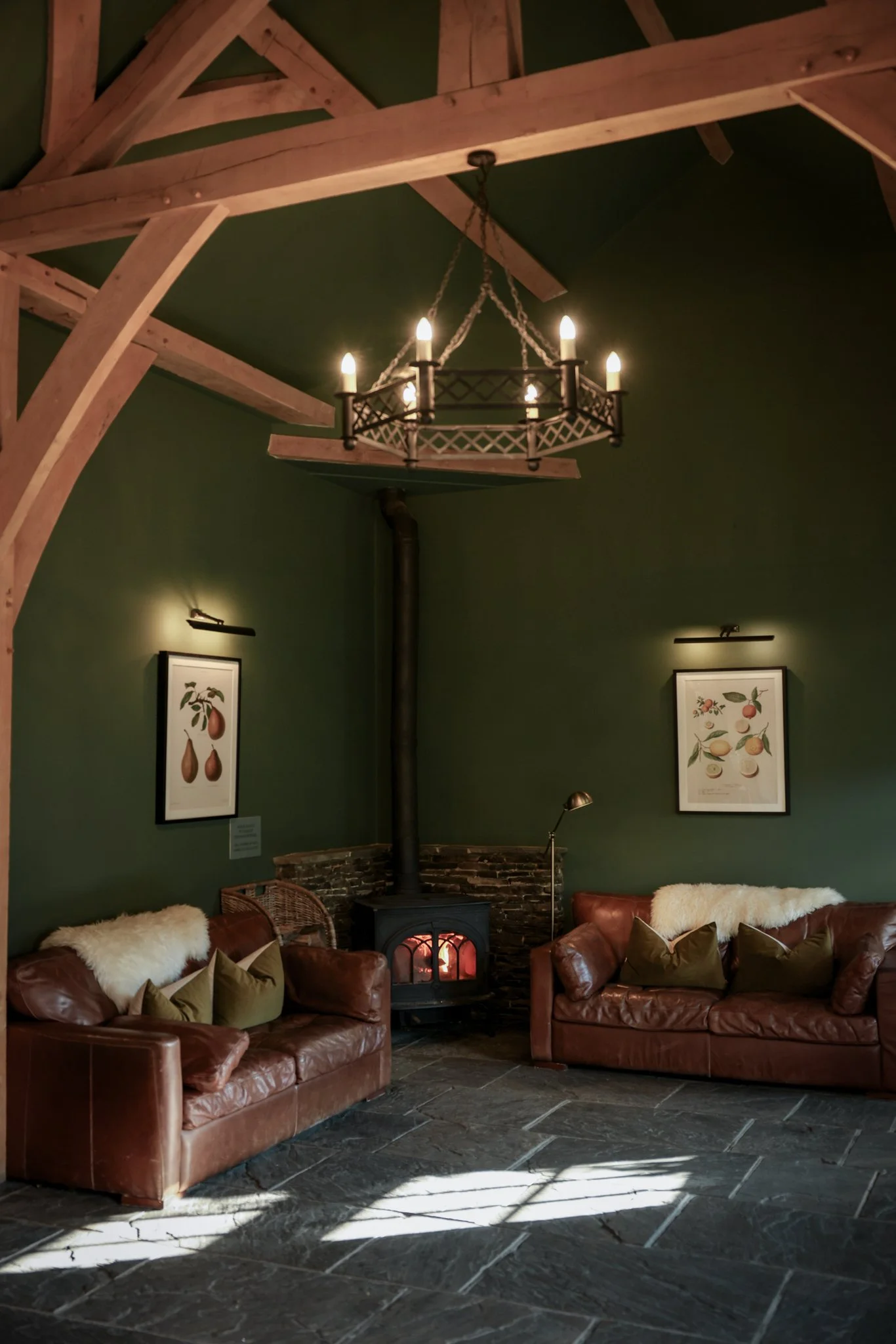 A cozy living room with two leather sofas, a wood stove, and framed botanical prints on dark green walls. A chandelier hangs from a wooden ceiling beam, casting warm light.