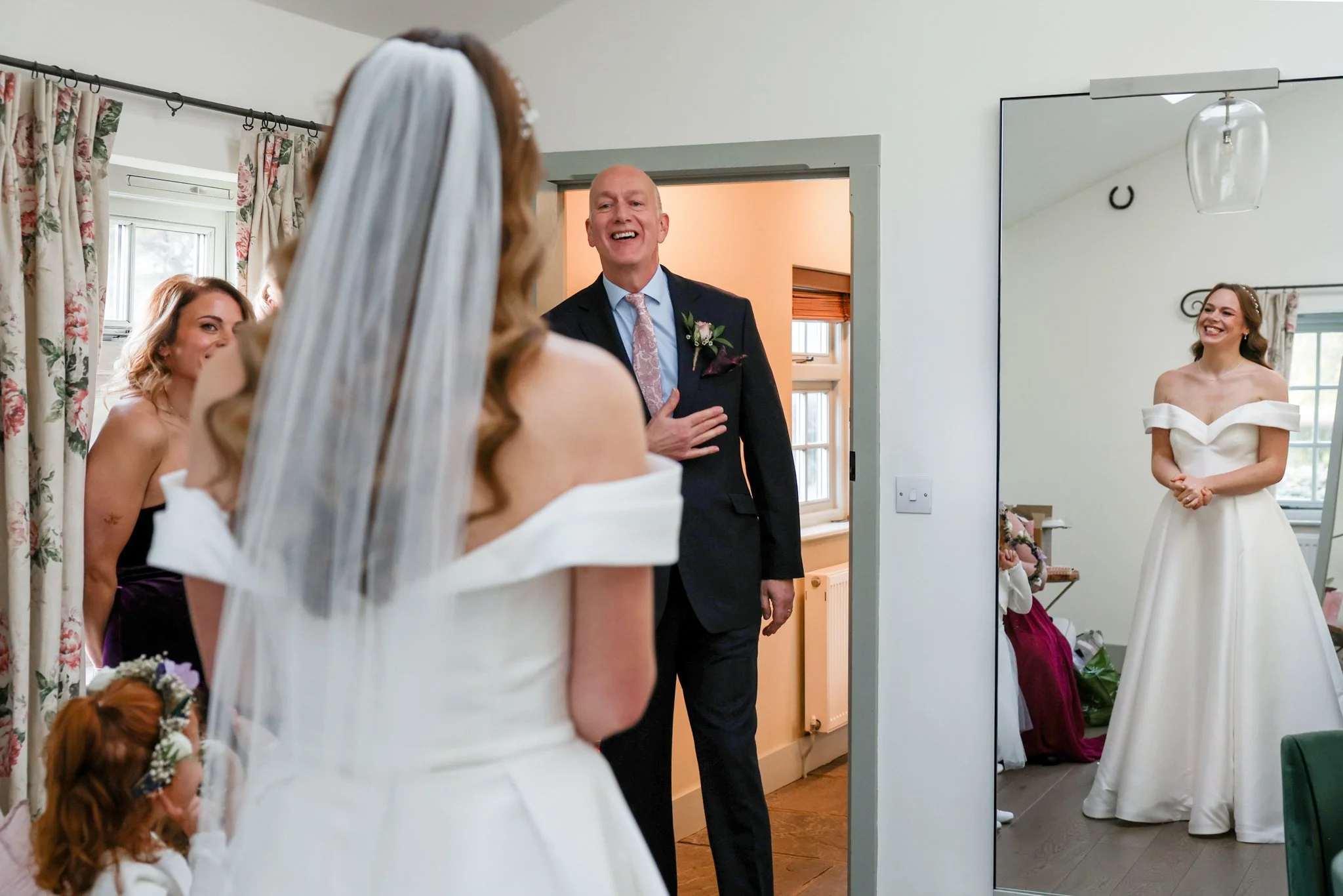 A wedding scene with a bride in a white dress and veil, two bridesmaids in purple dresses, and a groom in a suit with a boutonniere, smiling and standing near a mirror in a bright room.