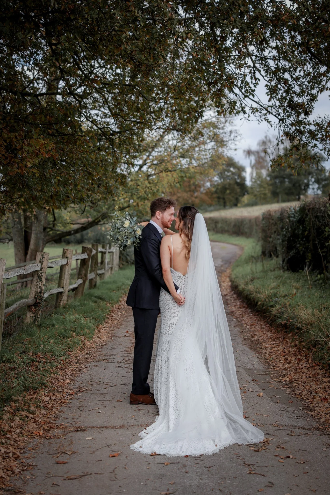 A bride and groom standing close together on a dirt path surrounded by trees and fallen leaves, holding hands and smiling at each other, outdoors during fall.