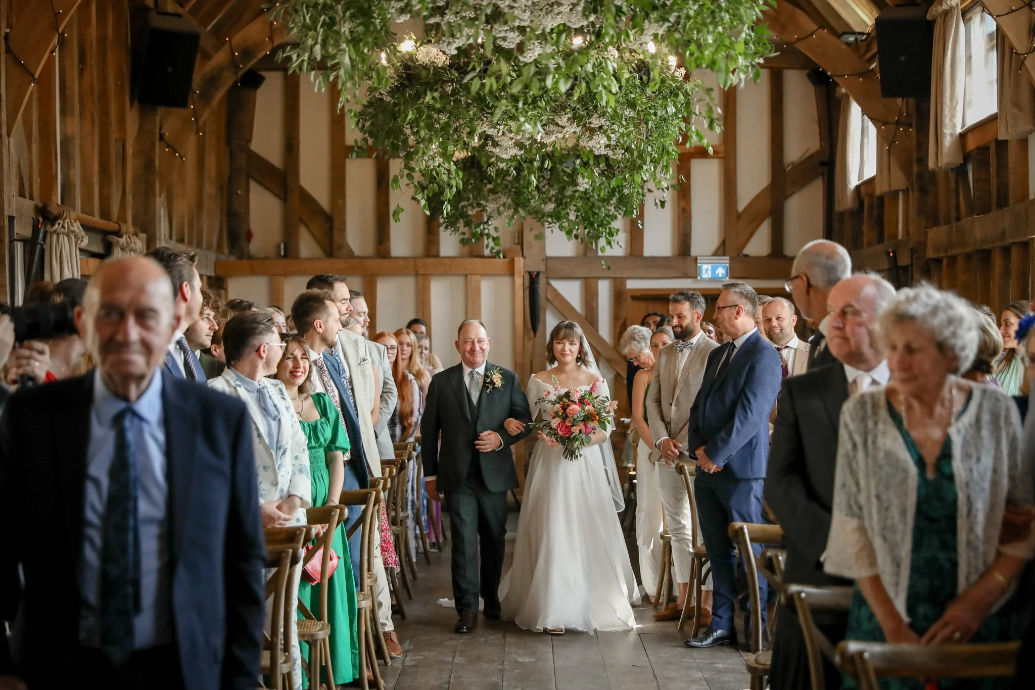 A bride and groom walking down the aisle at their wedding ceremony in a rustic barn with wooden beams and hanging greenery decoration, surrounded by guests.