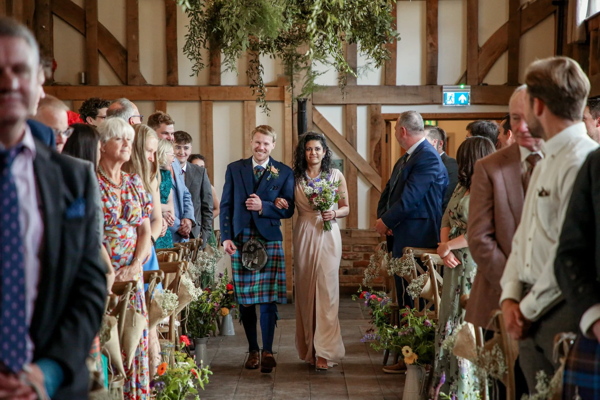 A bride and groom walking down the aisle at a wedding ceremony inside a rustic wooden venue, surrounded by seated guests and decorated with flowers and greenery.