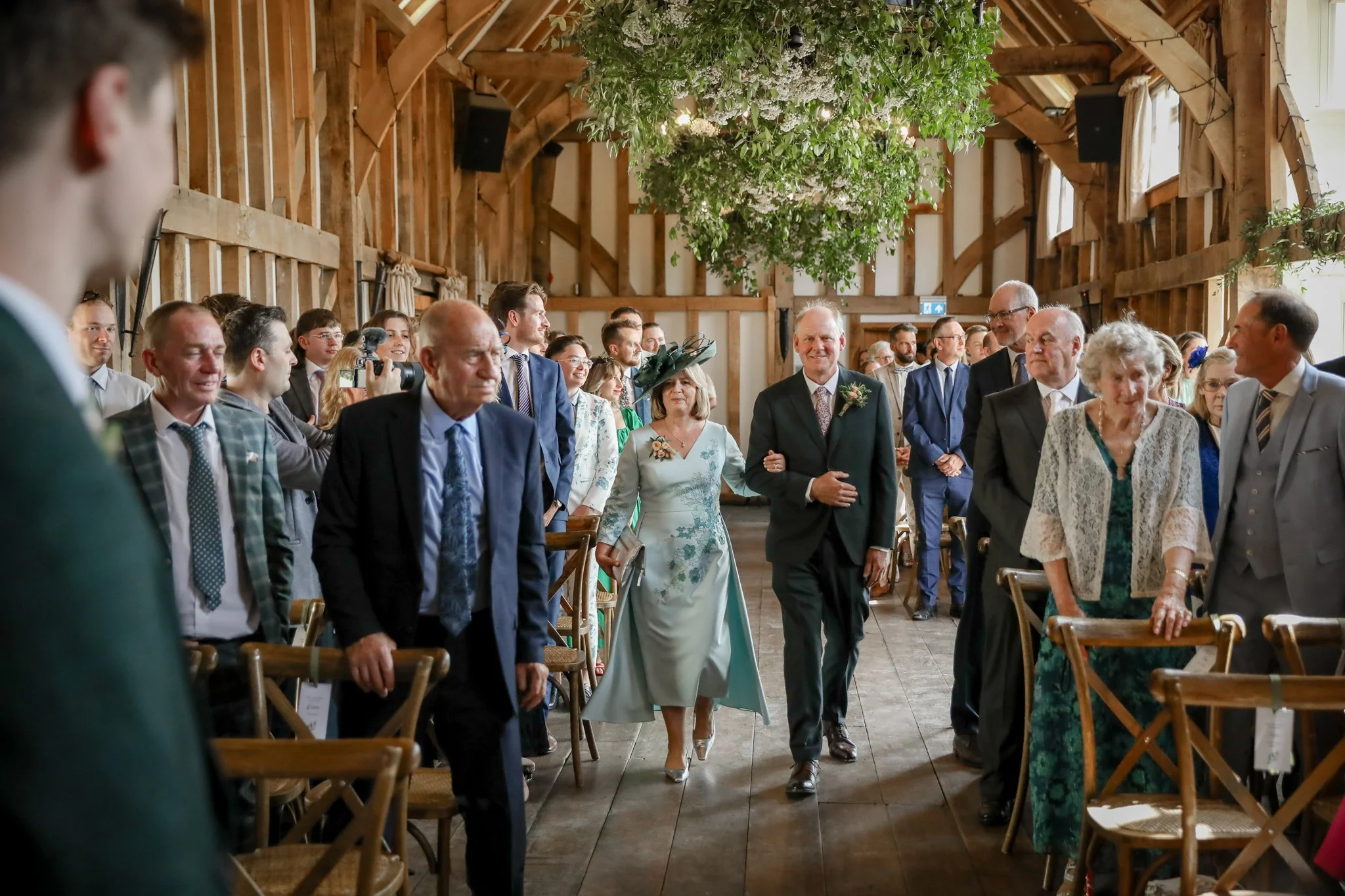 Guests at a wedding ceremony walking down the aisle inside a rustic wooden barn with green hanging plants from the ceiling.
