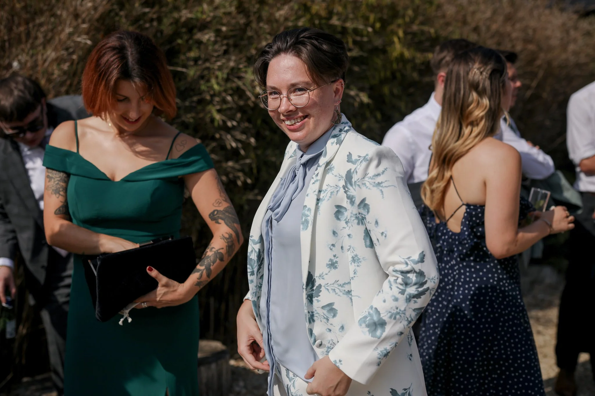 Group of people outdoors, with a woman with short dark hair, glasses, and a light floral blazer smiling at the camera in the foreground. Other individuals are engaging in activities or conversations behind her.