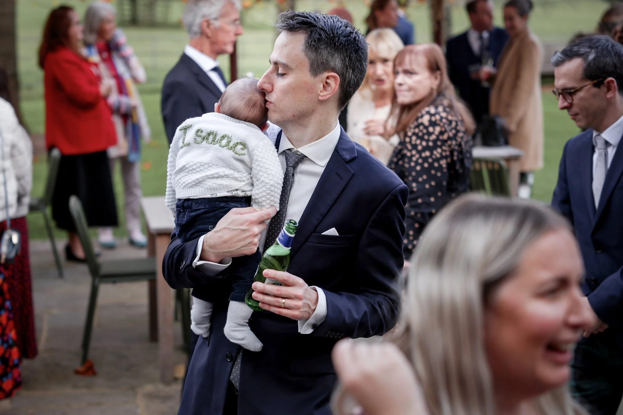 A man in a dark suit and tie is holding a small child who is wearing a white sweater that says 'I snack' and hugging him. The man is kissing the child's head while holding a beer bottle. Several people are in the background, standing and talking at a