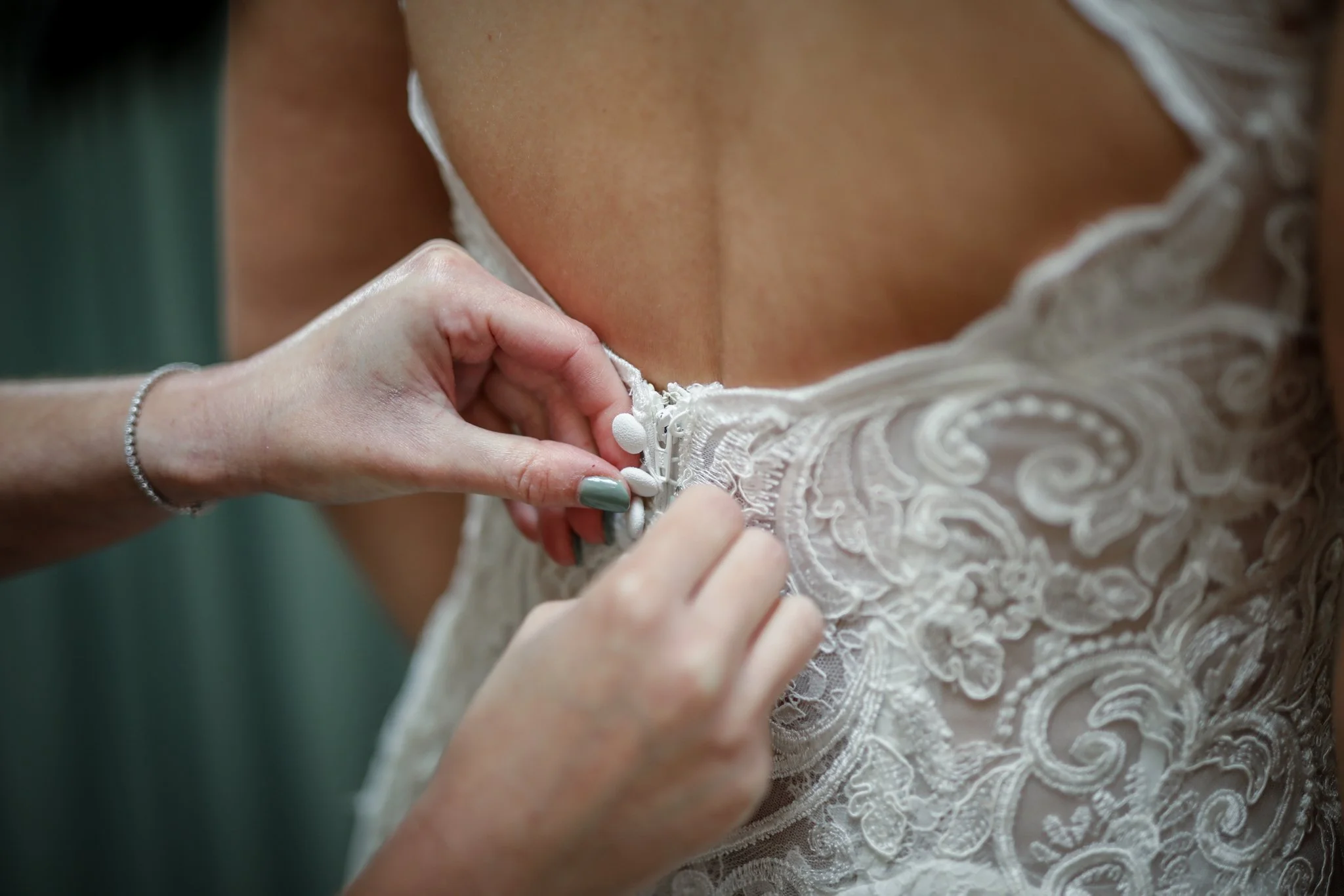 Close-up of a person fastening the zipper of a white lace wedding dress, with a silver bracelet on their wrist and painted nails.