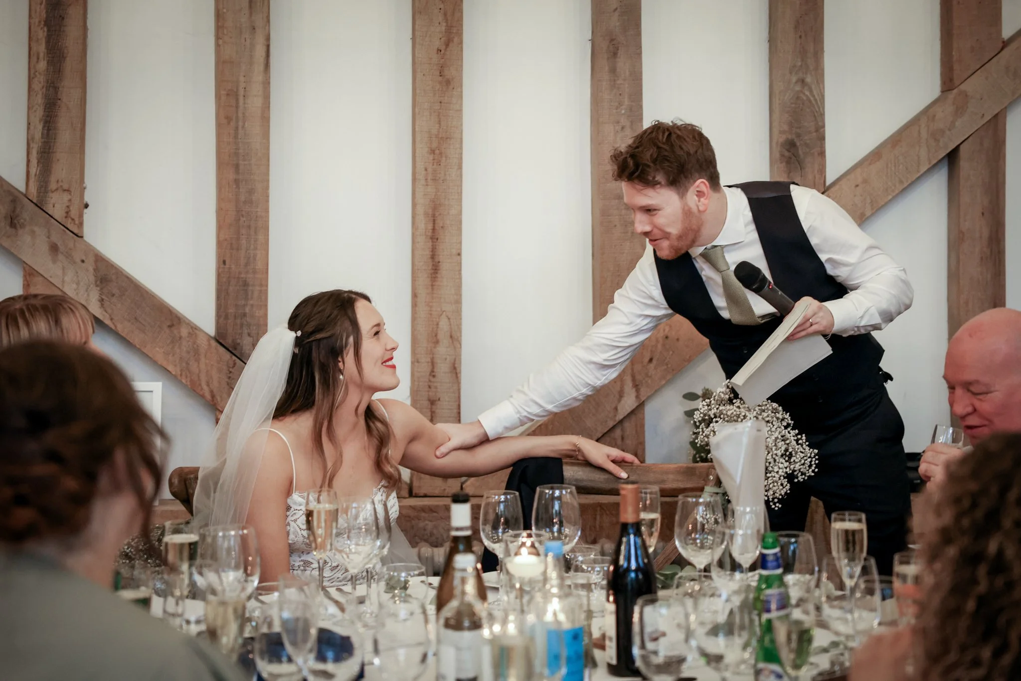 A groom in a vest and tie interacting with a bride who is wearing a wedding dress and veil at a wedding reception, with guests seated at tables and a rustic wooden wall in the background.