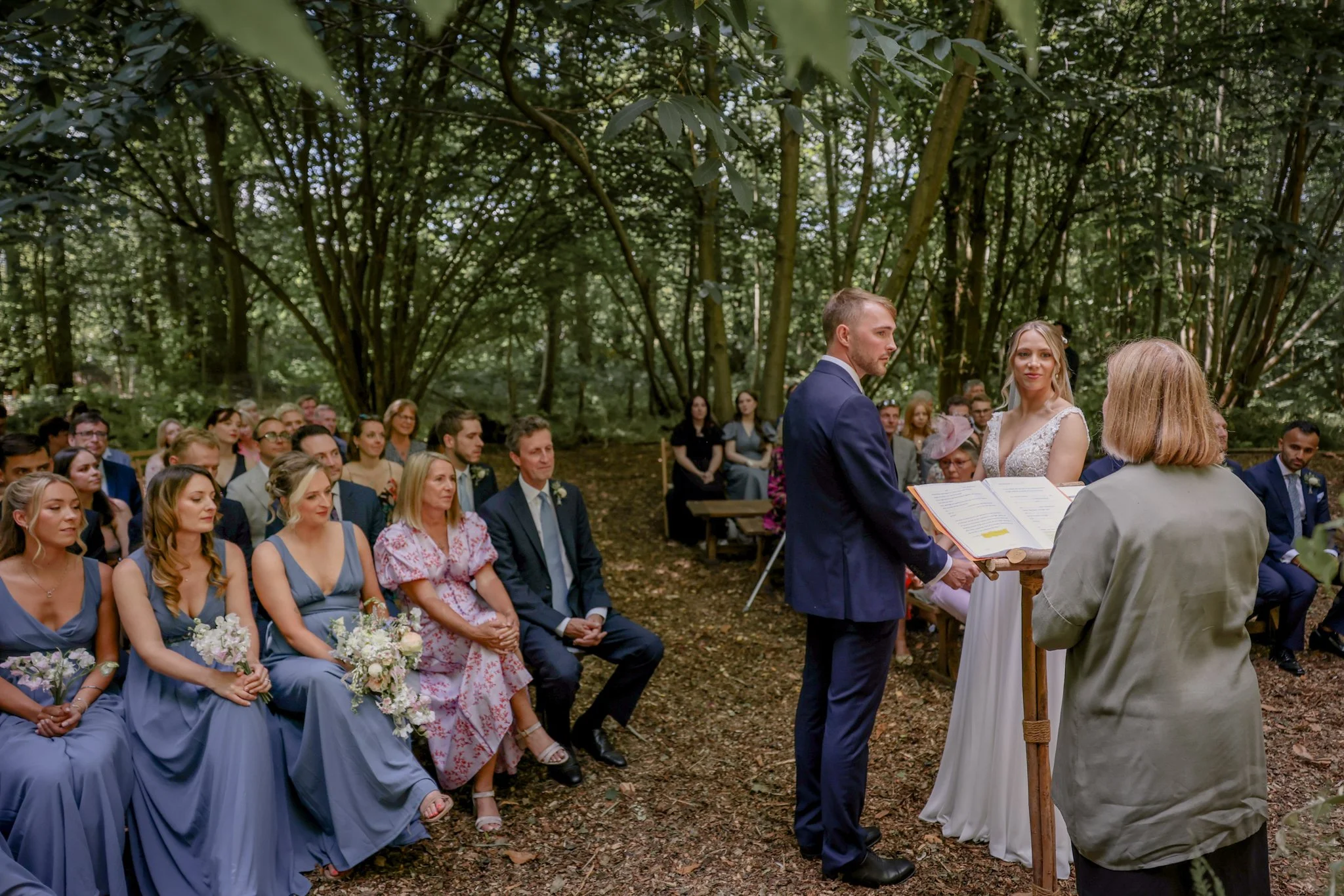 A wedding ceremony in a forest with a bride and groom standing facing each other, witnesses seated in blue dresses and suits, trees and leaves surrounding the scene.