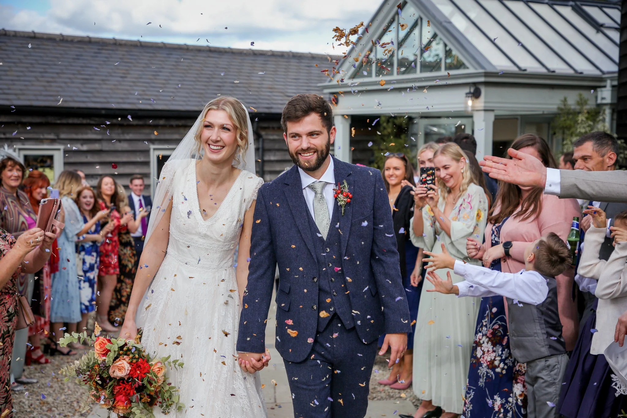 A newlywed couple holding hands and walking through a crowd of guests at an outdoor wedding, with confetti falling around them.