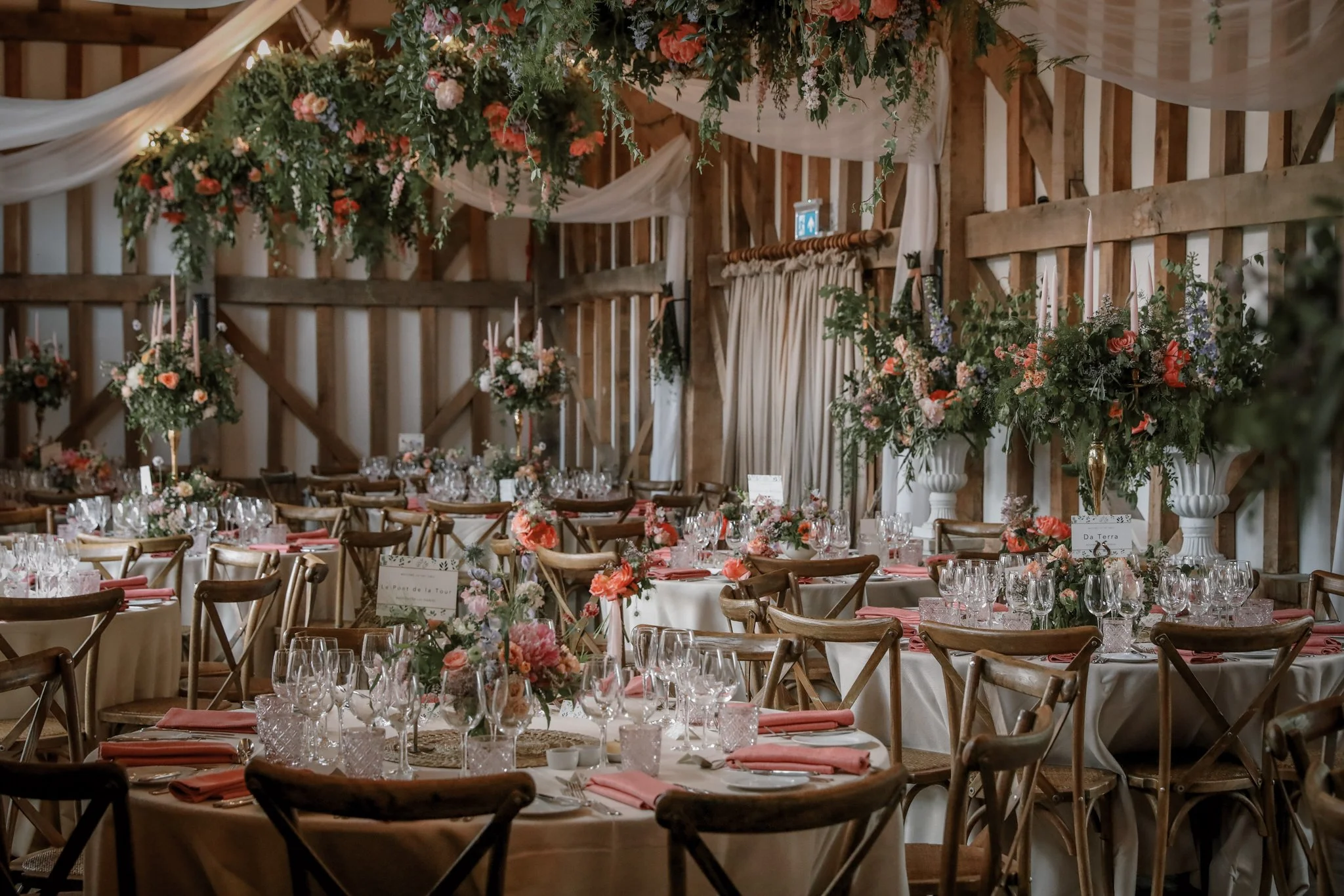 A decorated wedding reception hall with wooden walls, draped fabric, and floral arrangements on tables and hanging above the space.