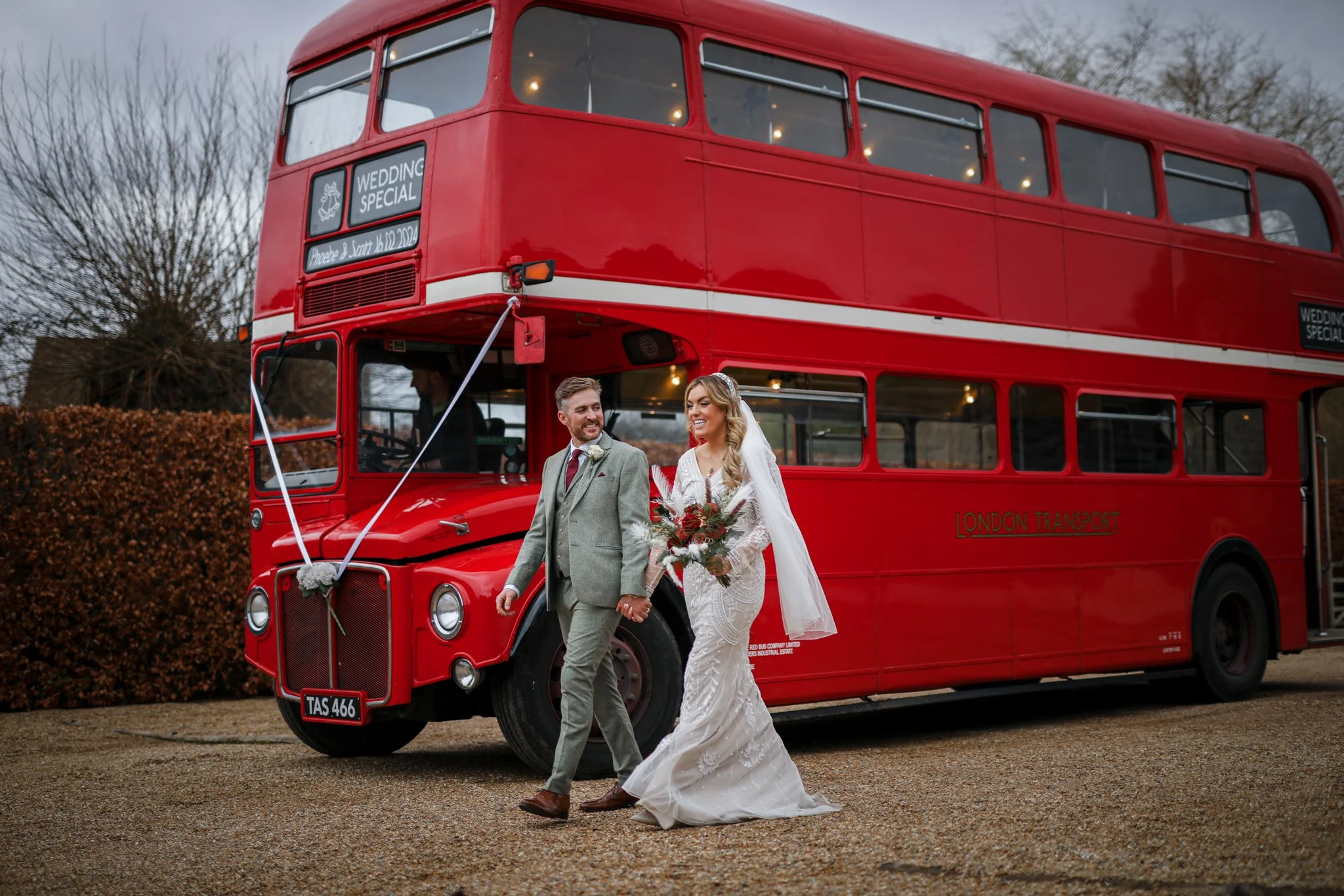 A bride and groom holding hands and walking in front of a red double-decker bus with a sign that says 'Wedding Special'. The bride is wearing a white wedding dress and veil, and the groom is dressed in a gray suit. The scene appears to be outdoor in 