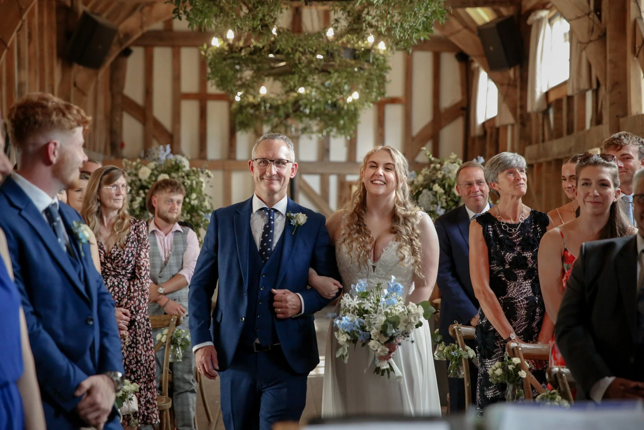 A wedding ceremony inside a rustic barn with the bride and groom standing together, surrounded by guests seated and standing, decorated with flowers and greenery, with warm lighting and wooden beams overhead.