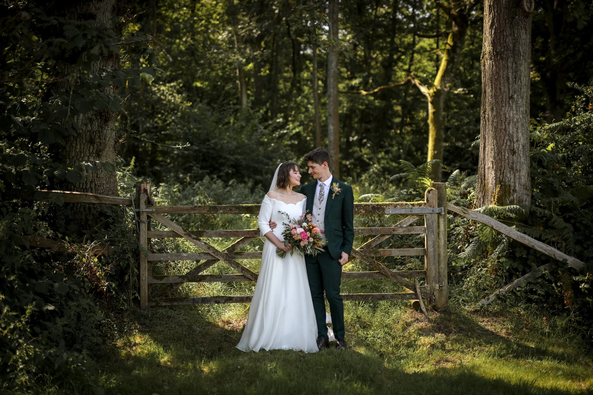 A bride and groom standing together outdoors near a wooden fence in a forested area, smiling at each other, with sunlight filtering through the trees.