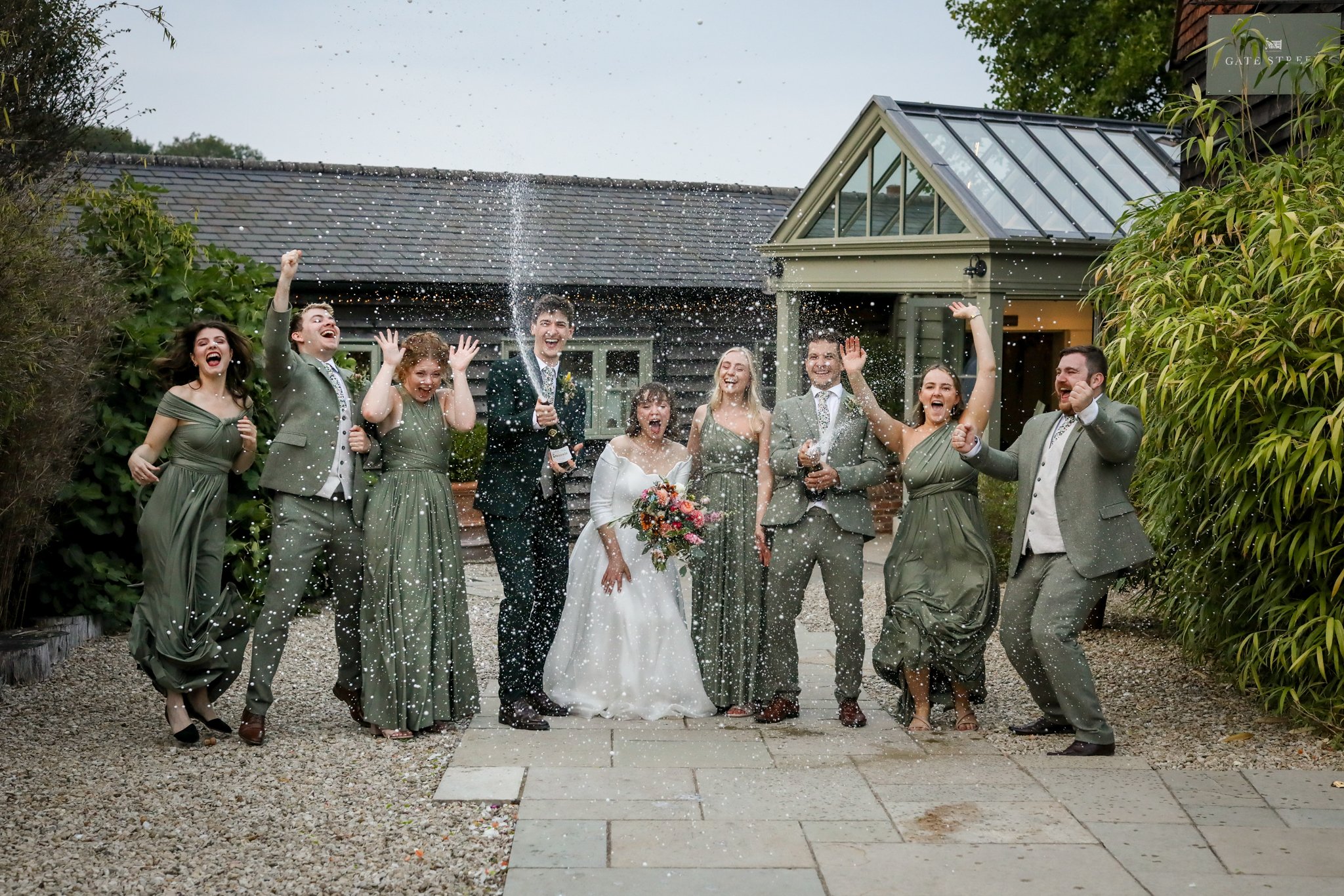 Group of people celebrating a wedding outdoors, throwing confetti, with the bride holding a bouquet and wearing a white dress, and the groom in a suit.