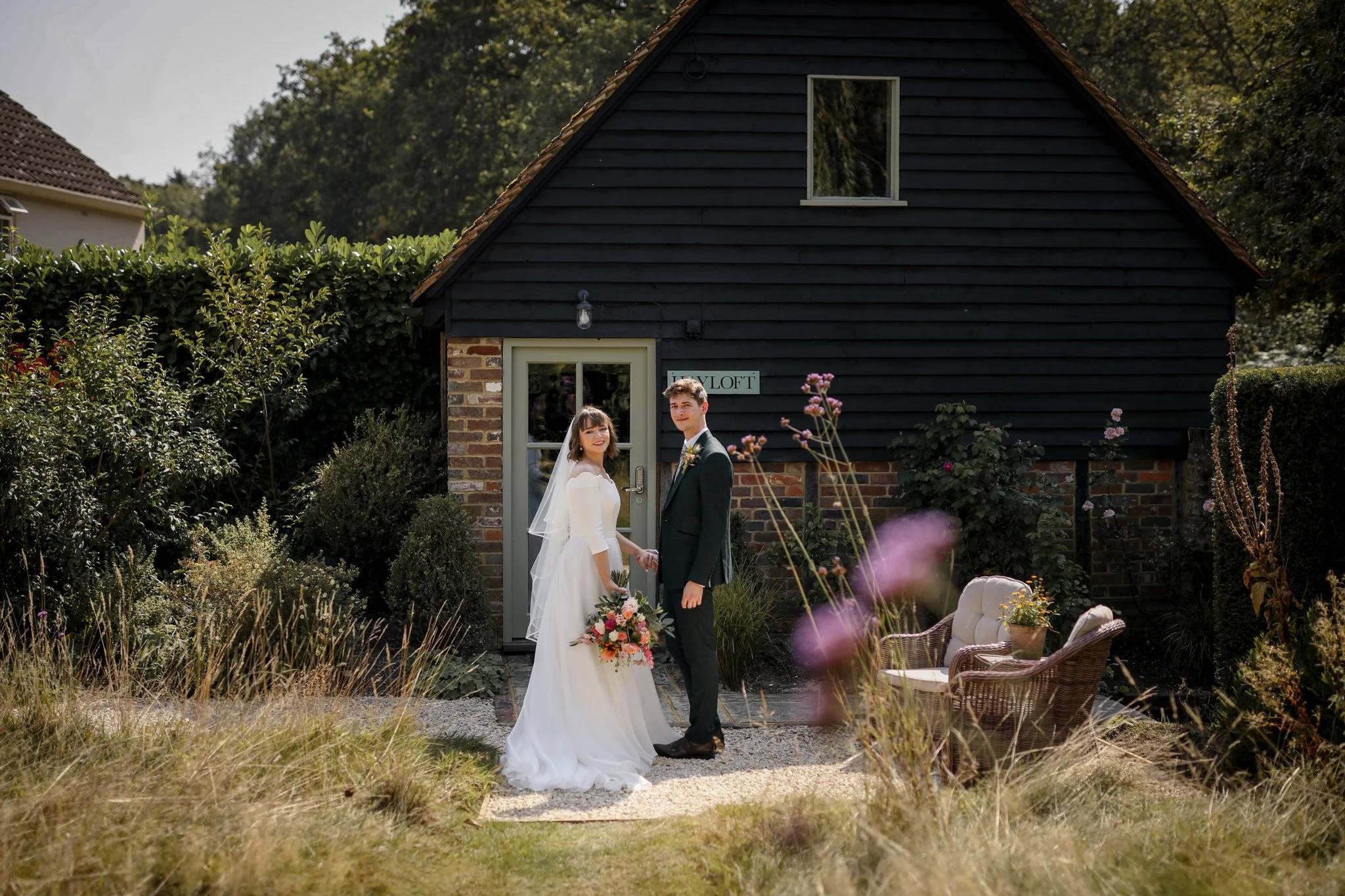 A bride and groom stand hand in hand in a garden outside a dark-colored house with a sign that reads 'YLOFT'. The bride wears a white wedding dress and veil, holding a bouquet, while the groom wears a dark suit. There are lush green plants and flower