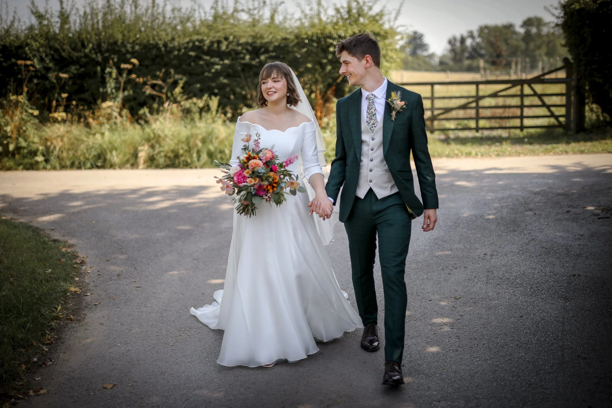 A bride and groom walking hand in hand outdoors on a sunny day. The bride is smiling, holding a colorful bouquet, and wearing a white wedding dress with an off-the-shoulder neckline. The groom is dressed in a dark green suit with a patterned tie and 