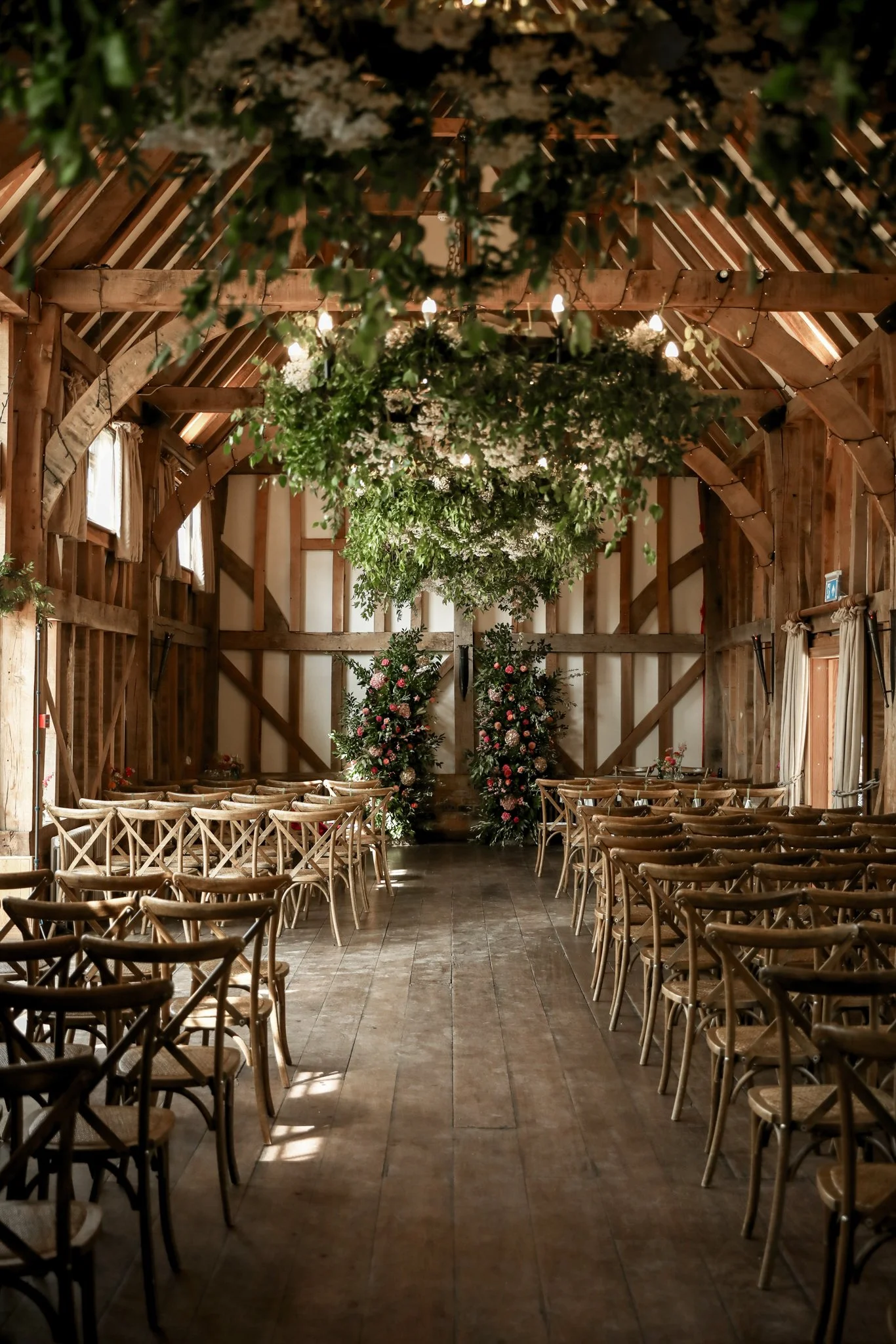 Interior of a rustic wooden barn decorated for a wedding ceremony, featuring hanging greenery and white flowers, with wooden chairs arranged in rows on either side of an aisle.