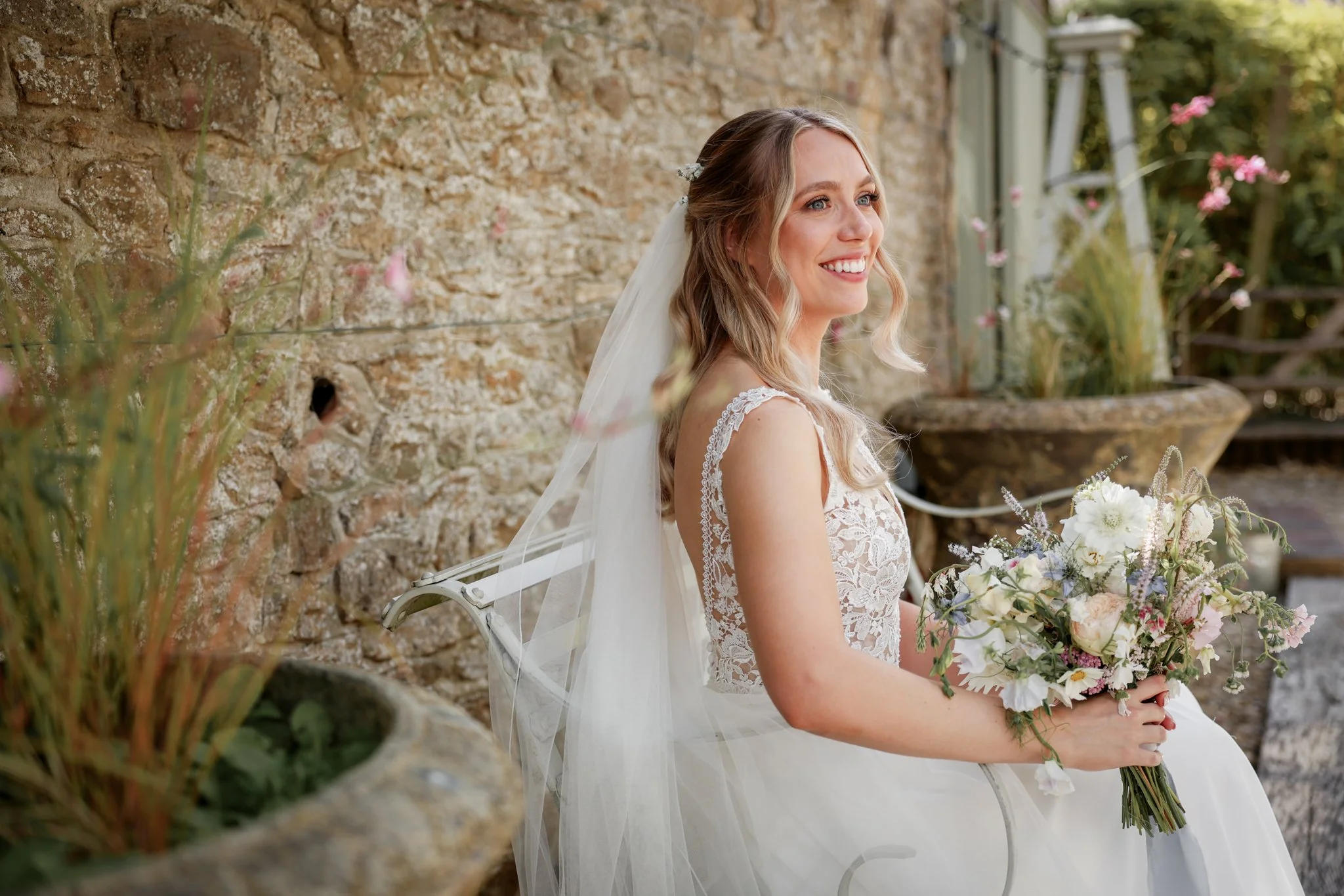A smiling bride with blonde hair in loose waves, wearing a lace wedding gown with a sheer back, holding a bouquet of flowers, standing outdoors near a stone wall with greenery and pink flowers.