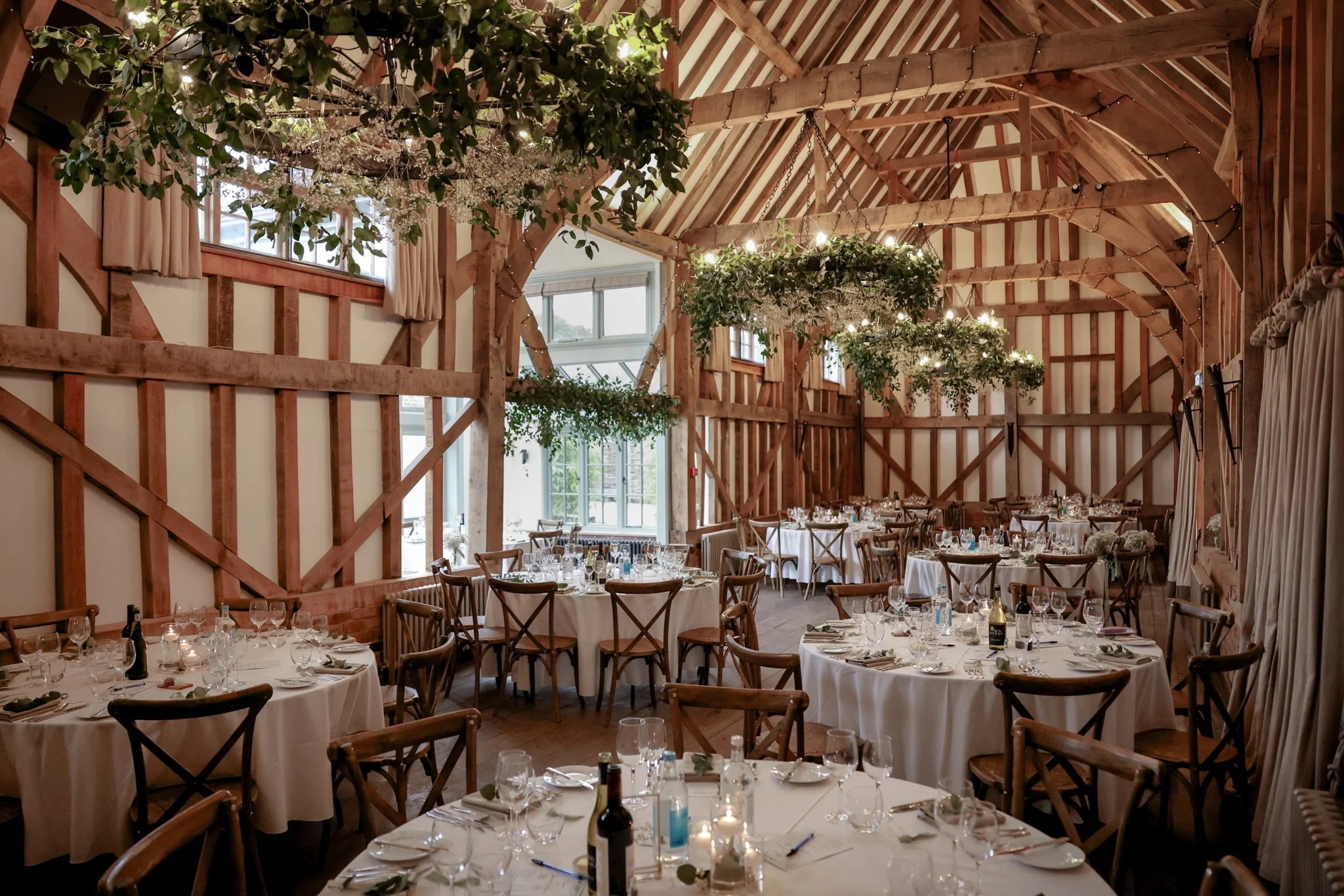 A large banquet hall decorated for an event with round tables covered in white tablecloths, set with glassware, plates, and utensils. The hall has wooden beams, hanging greenery decorations, and string lights. There are large windows with curtains al