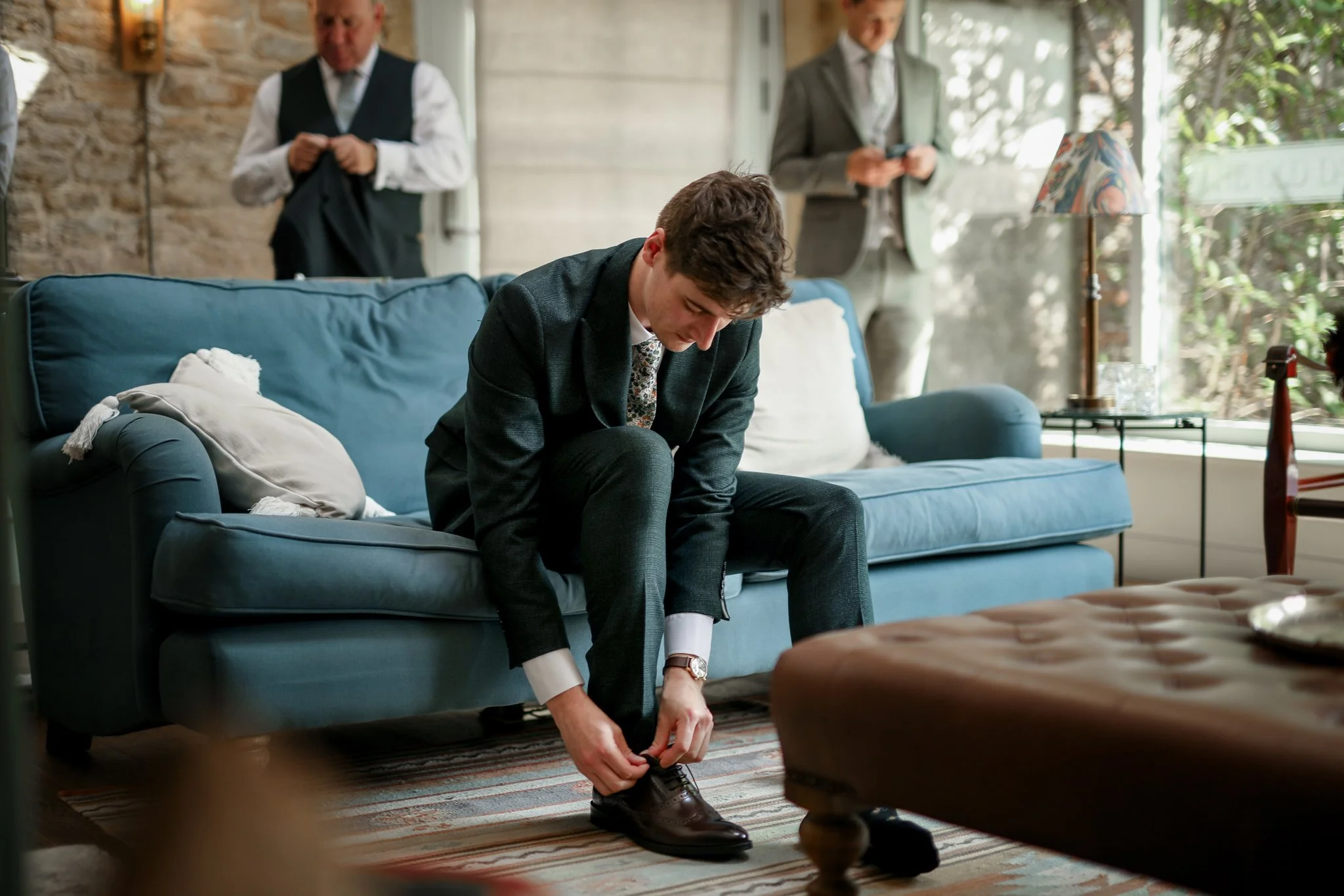 A young man in a suit is sitting on a blue couch, tying his shoe.