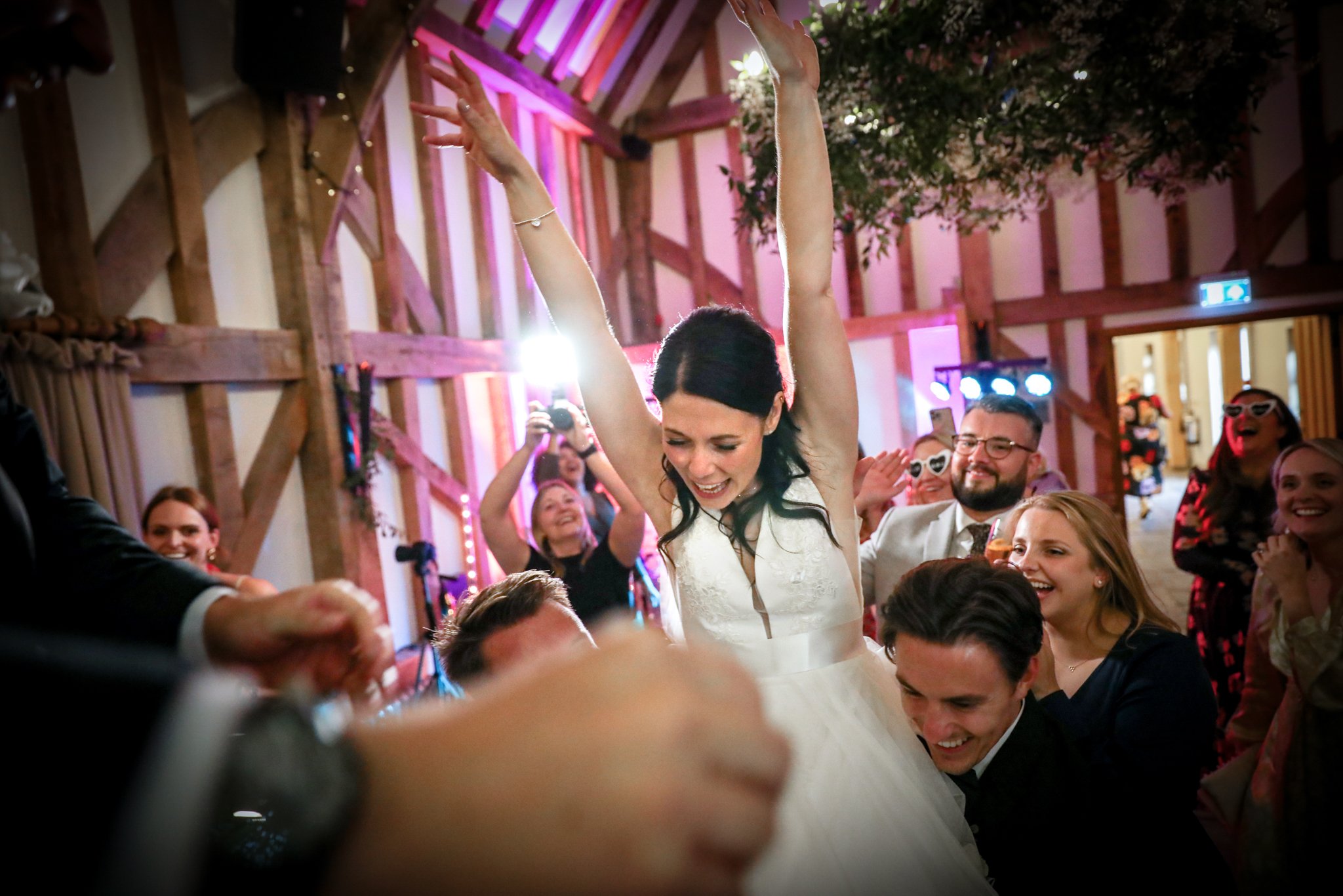A bride in a white wedding dress raises her arms with joy during her wedding reception, surrounded by smiling guests in a rustic venue decorated with warm lighting and wooden beams.