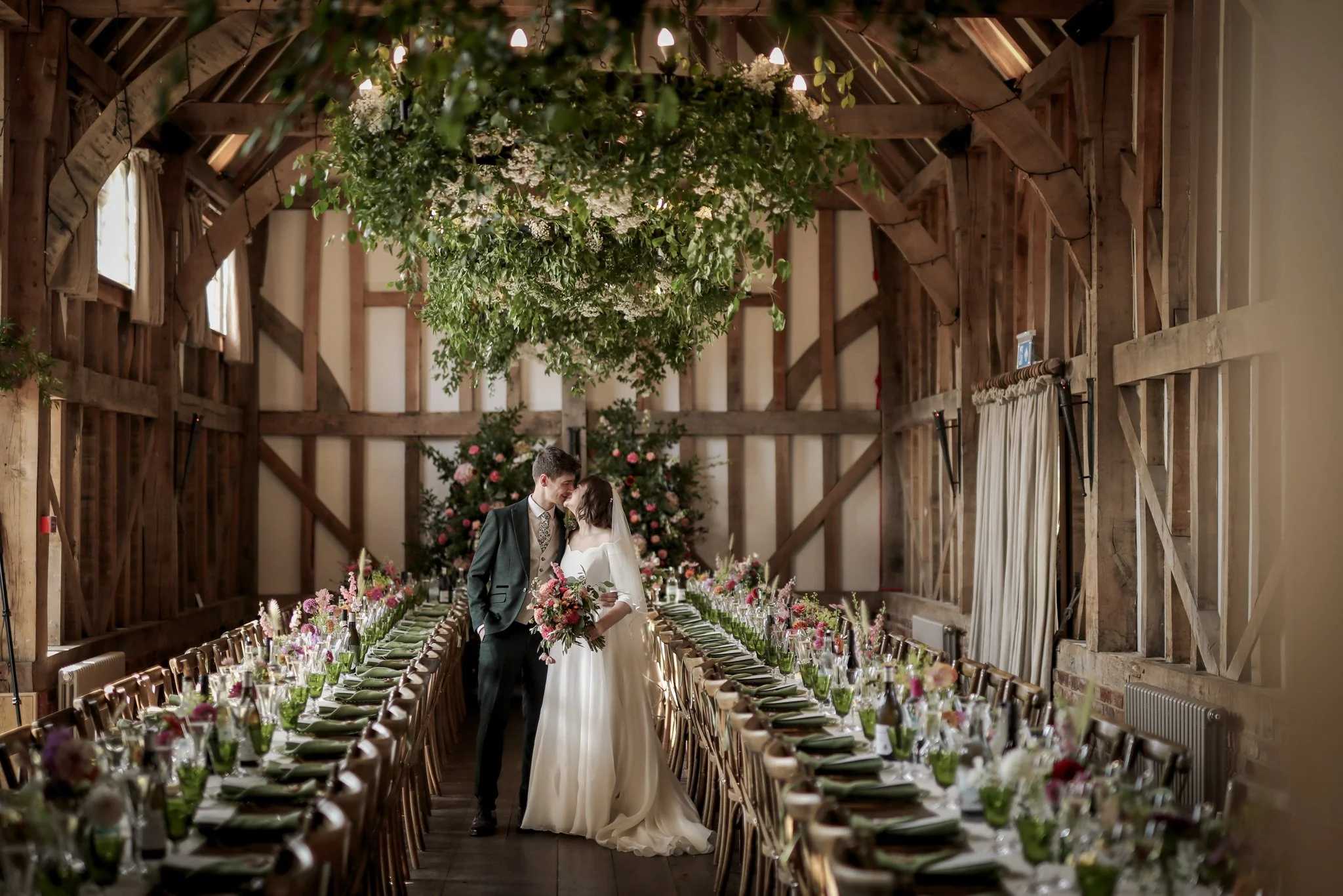 A bride and groom are kissing inside a rustic barn decorated for a wedding reception, with long banquet tables set with flowers and place settings.