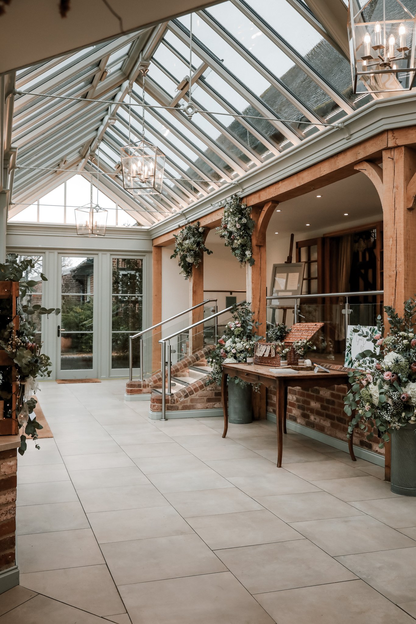 Indoor space with a glass ceiling, hanging chandeliers, wooden beams, floral decorations, and a table with a basket and small items.