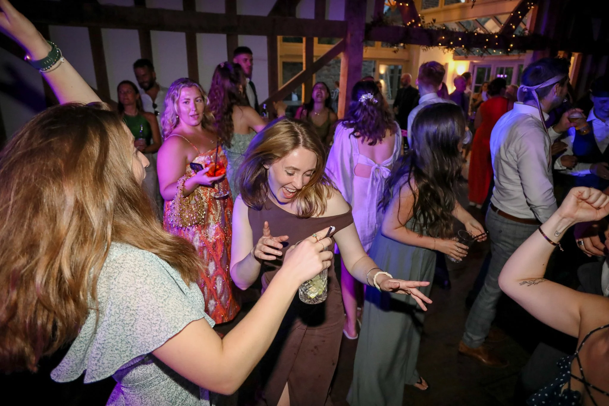 A group of people dancing and socializing at a party or event in a dimly lit indoor venue with string lights and wooden beams.