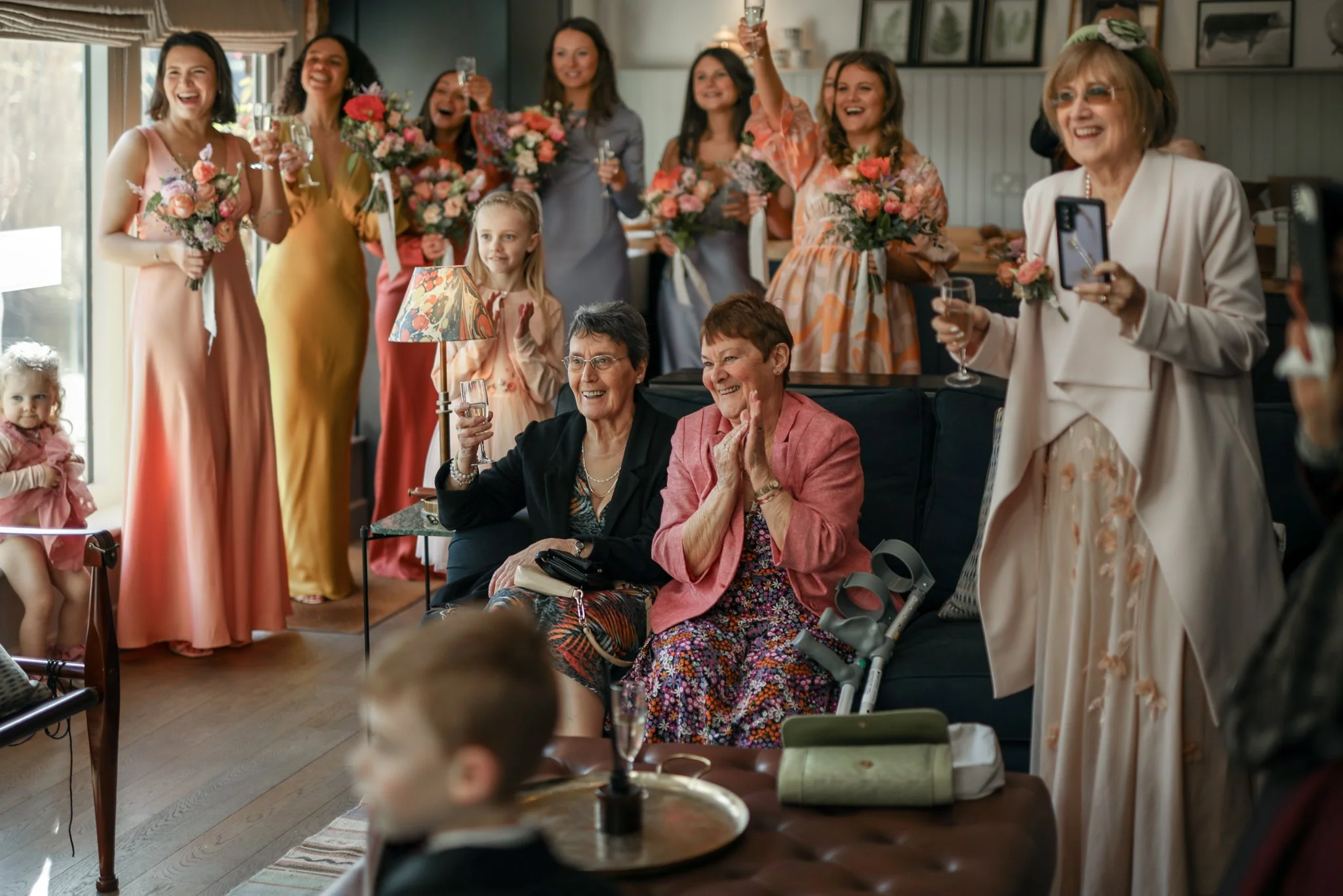 A group of women, including older women and younger women, and two young girls celebrating with champagne and flowers in a cozy, decorated indoor setting, possibly at a wedding or special event.