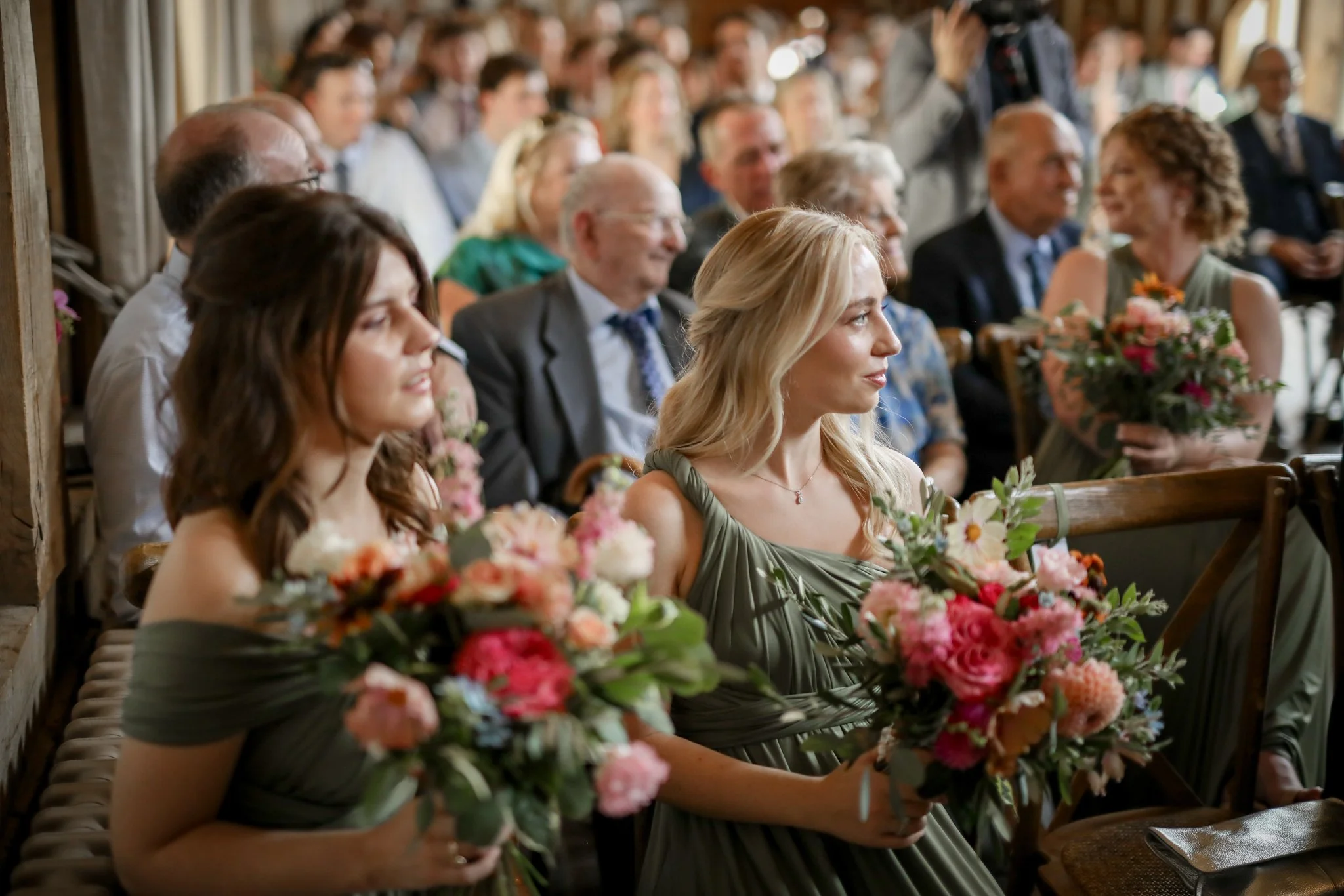 Guests seated at a wedding reception, holding bouquets of pink and white flowers, with a woman in a green dress in the foreground.
