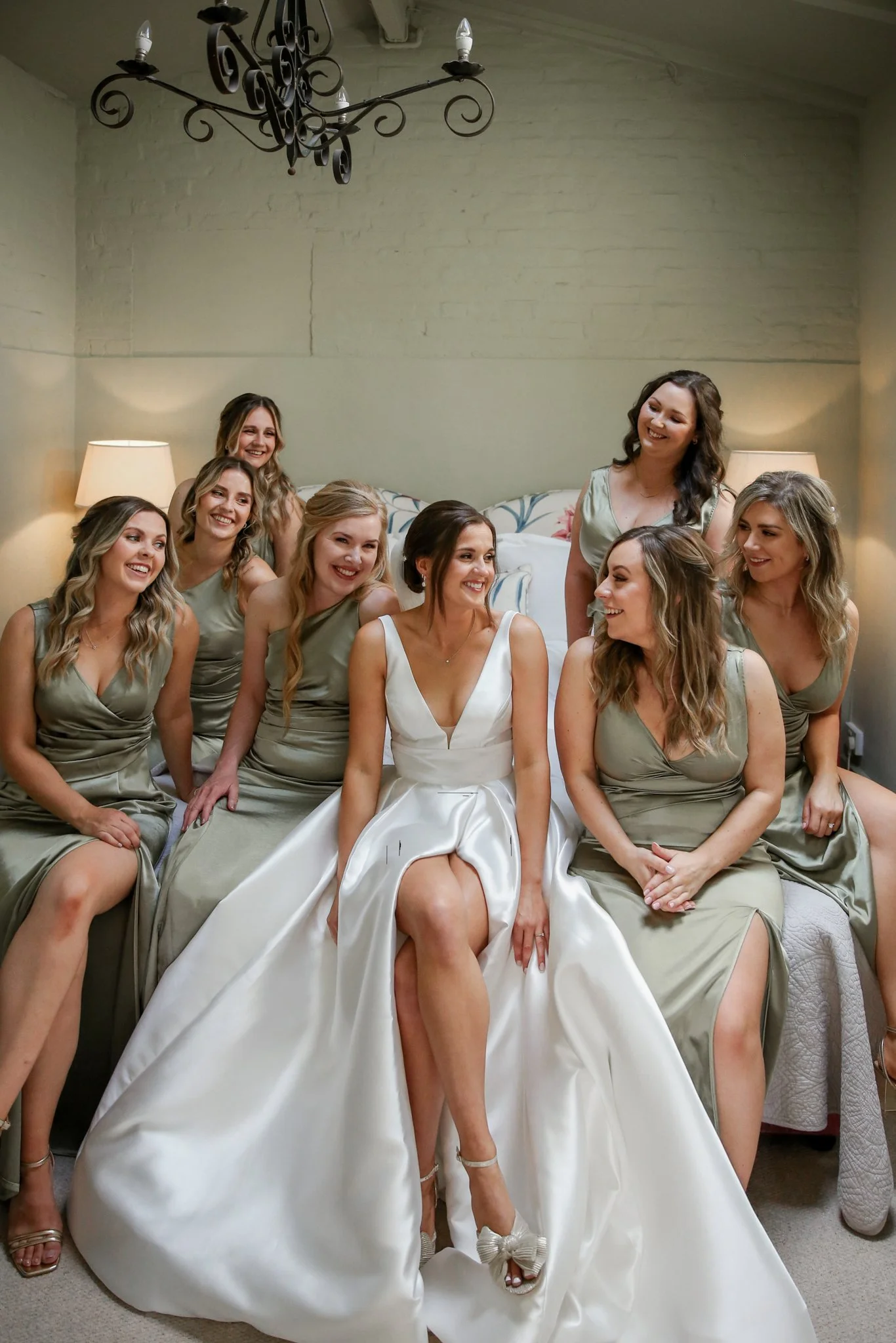 A bride and seven bridesmaids sitting on a bed in a room with beige walls and a chandelier. The bride is in a white wedding dress, and the bridesmaids are in matching light green dresses. They are all smiling and laughing.