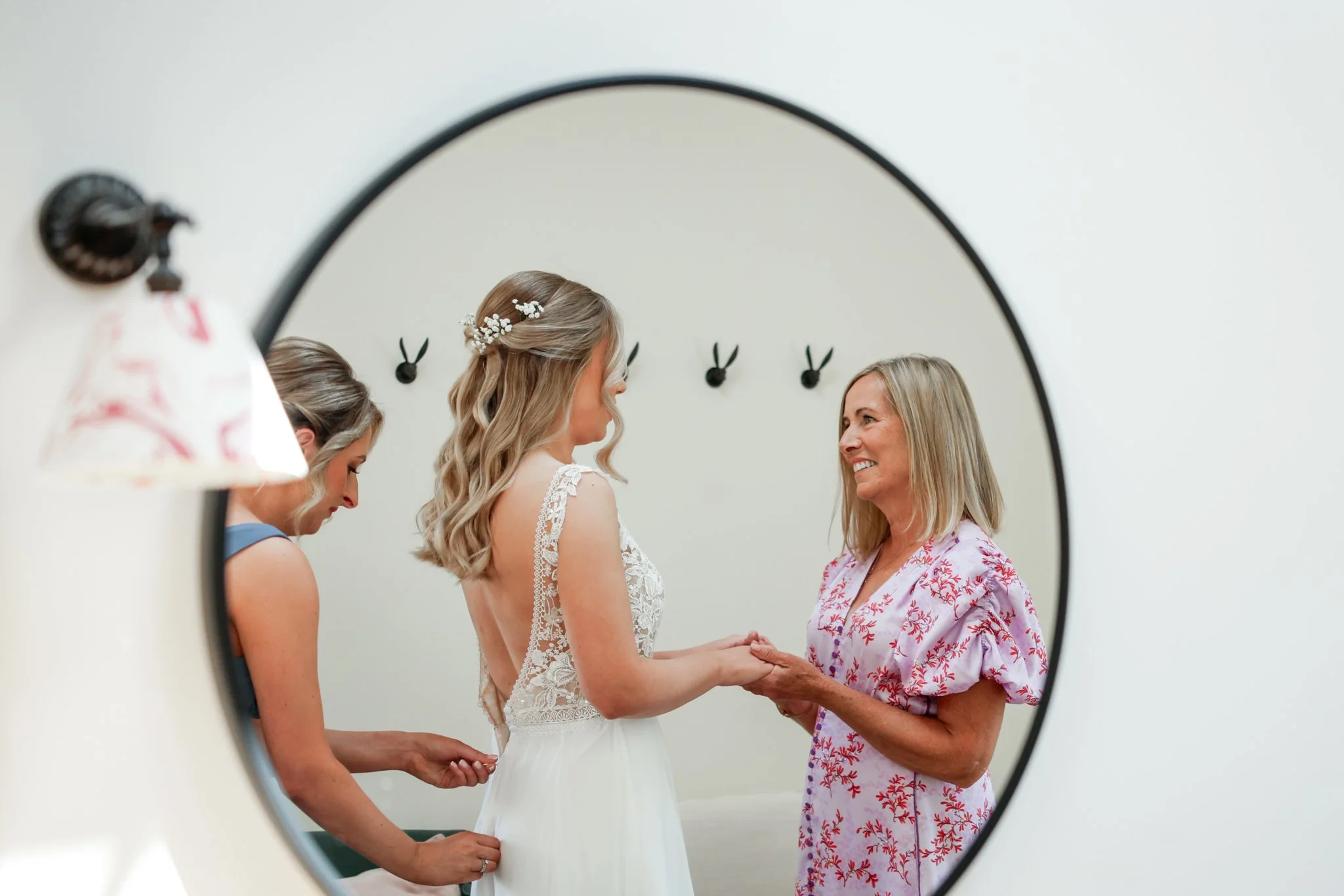 A bride holding hands with a woman, possibly her mother, during a wedding preparation session, as seen through a mirror.