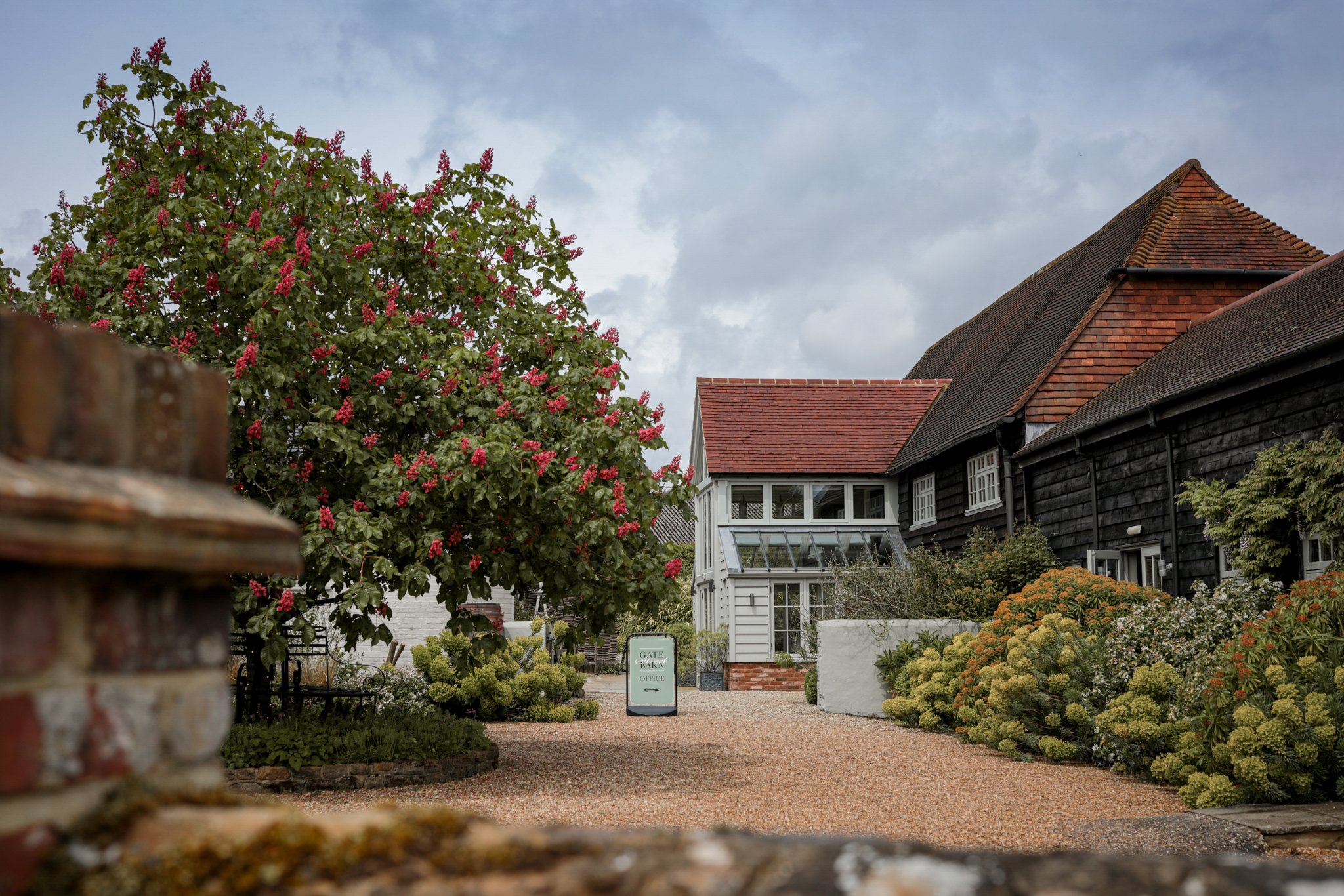 A garden scene with lush green bushes and trees with pink flowers, a gravel pathway, and a house with dark wooden siding and red-tiled roofs under a cloudy sky.