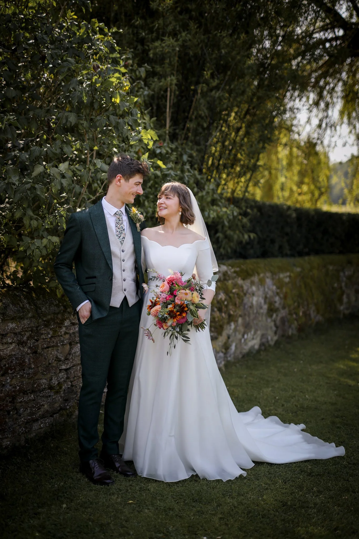 A wedding couple stands outdoors near a stone wall and greenery, smiling at each other. The bride holds a bouquet of flowers, wearing a white wedding dress with an off-the-shoulder neckline and veil. The groom wears a dark suit with a patterned tie, 