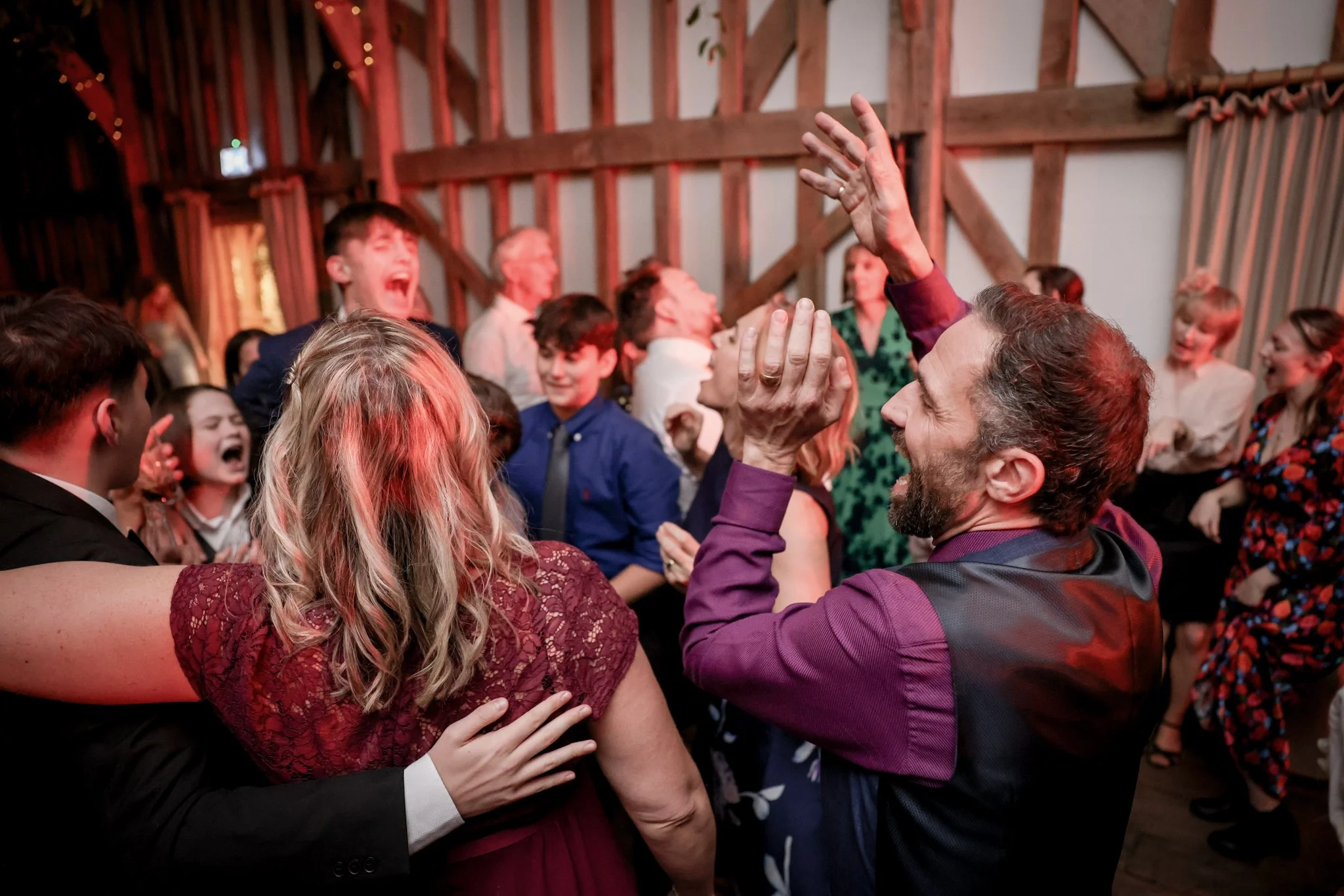 People dancing and celebrating at a lively party or wedding reception inside a rustic barn, with warm lighting and wooden beams.