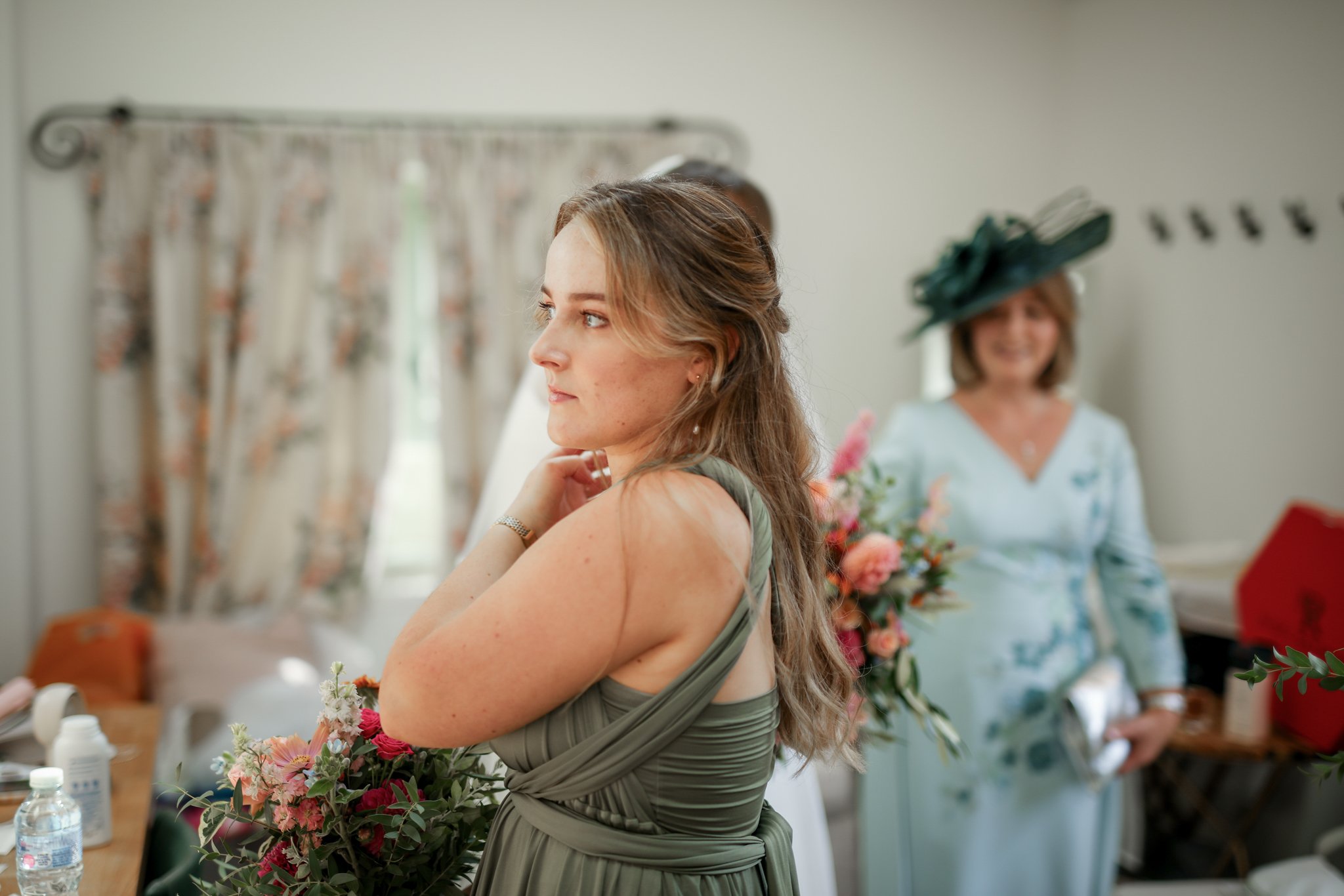 A woman with long wavy hair and a light dress stands in front of a mirror, adjusting her collar during a wedding preparation. Behind her, another woman with a large hat and floral outfit holds a bouquet. The room has light-colored walls and a table w
