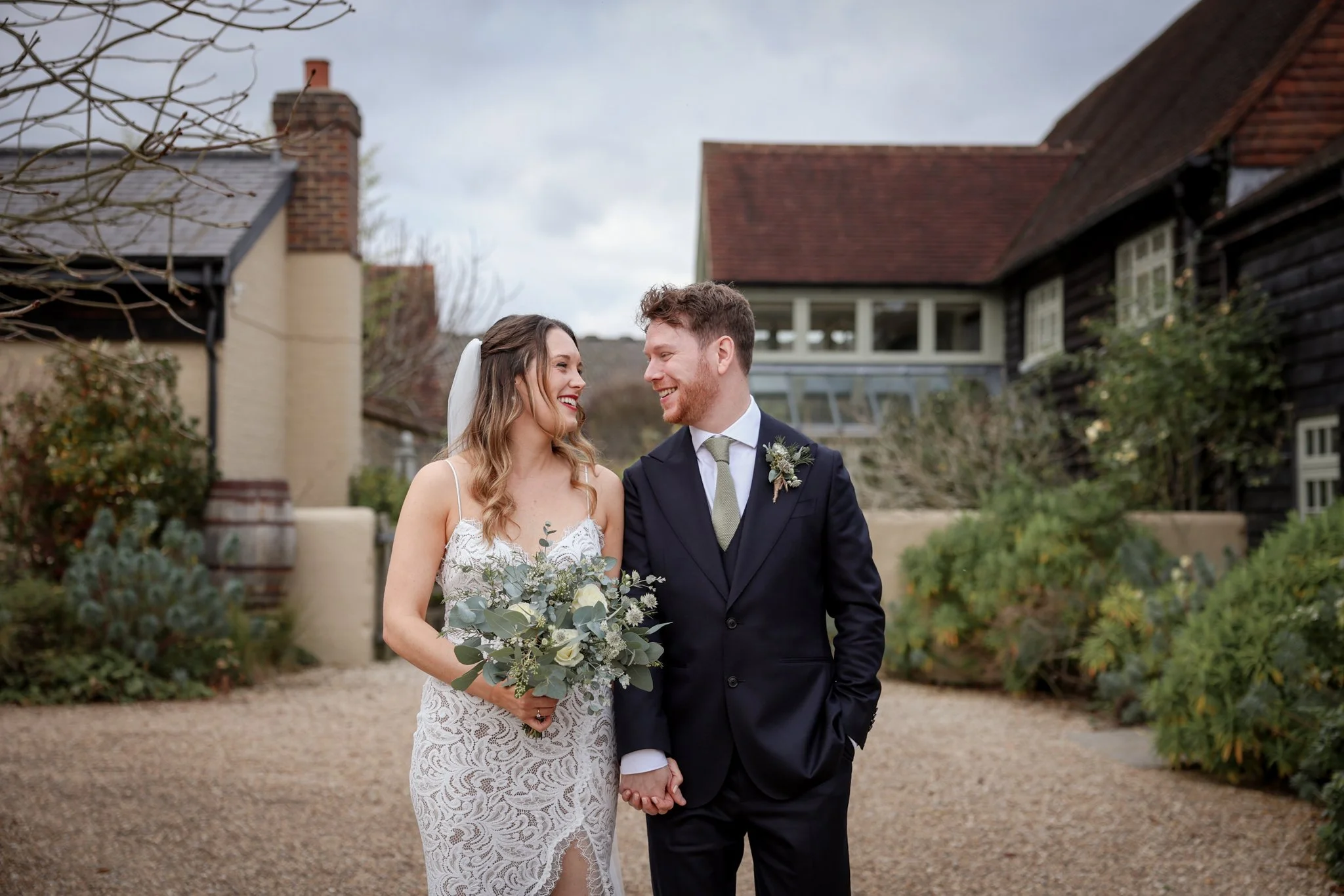 A bride and groom holding hands and smiling at each other outdoors, with houses and trees in the background, on a cloudy day.