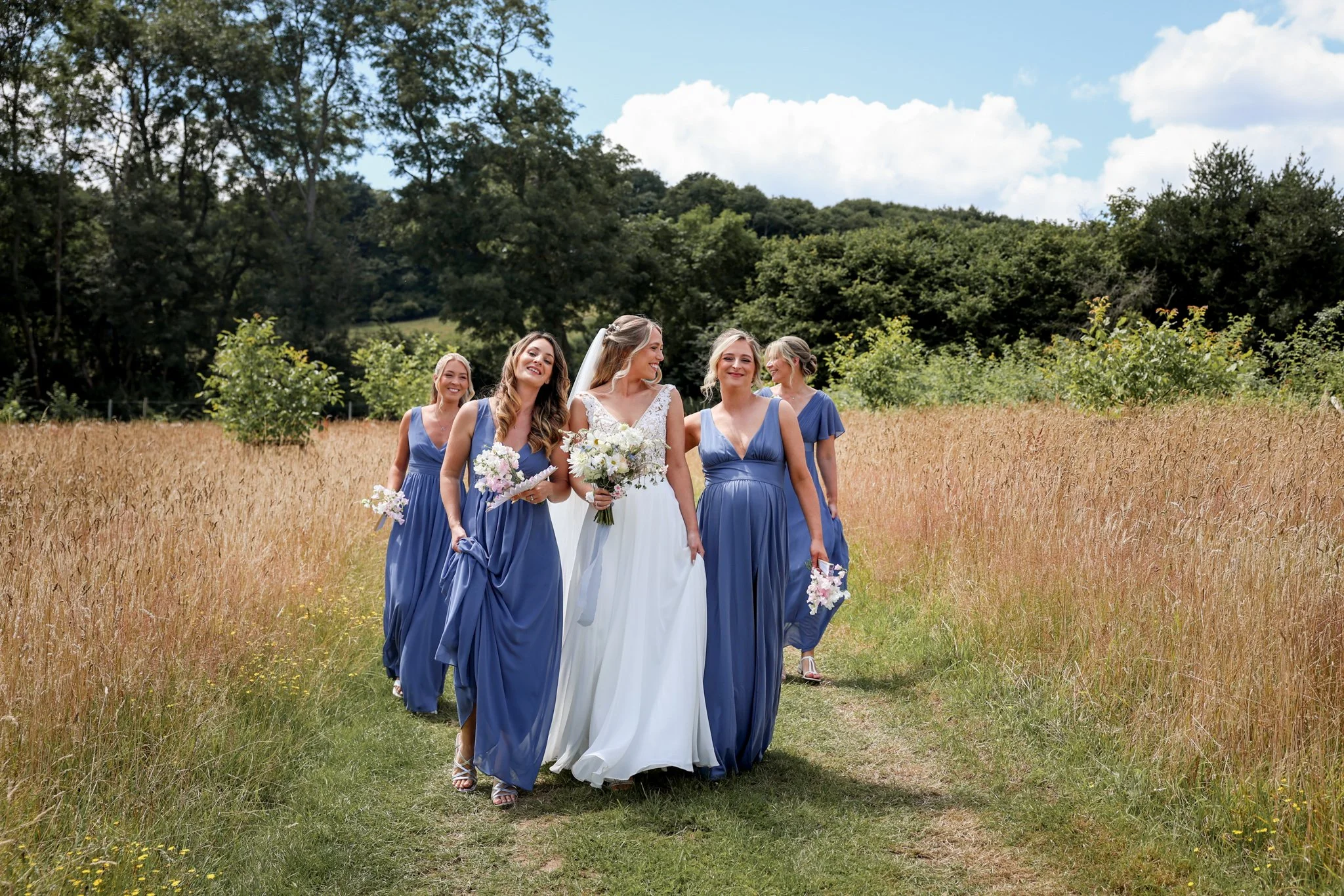 A bride in a white wedding dress and five bridesmaids in blue dresses walking through a field with trees in the background.