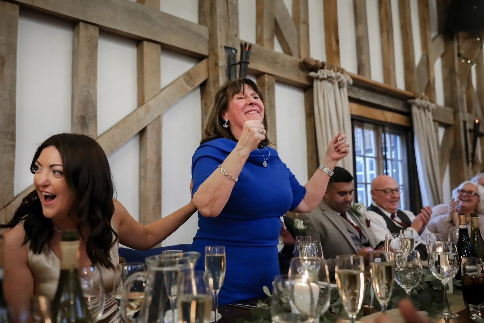 A woman in a blue dress celebrating at a dinner party, smiling and holding hands with others around the table in a rustic, warmly lit setting.