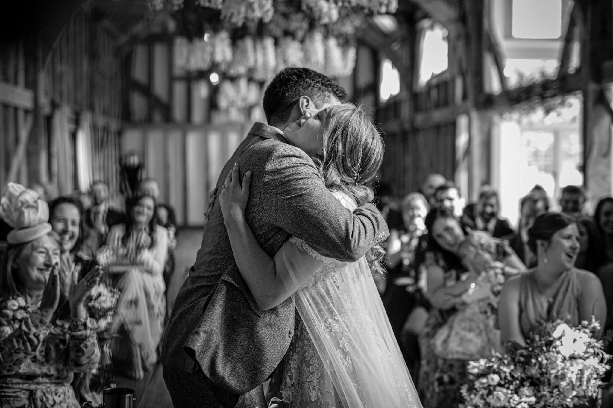 A black and white photo of a wedding couple sharing a kiss during their ceremony, with guests seated and watching in the background.