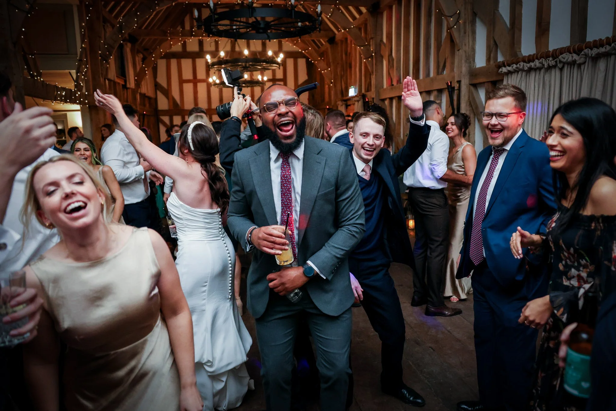A lively wedding reception in a rustic barn with string lights and chandeliers, showing happy guests dancing and celebrating, including a groom holding a drink and smiling, surrounded by friends and family.