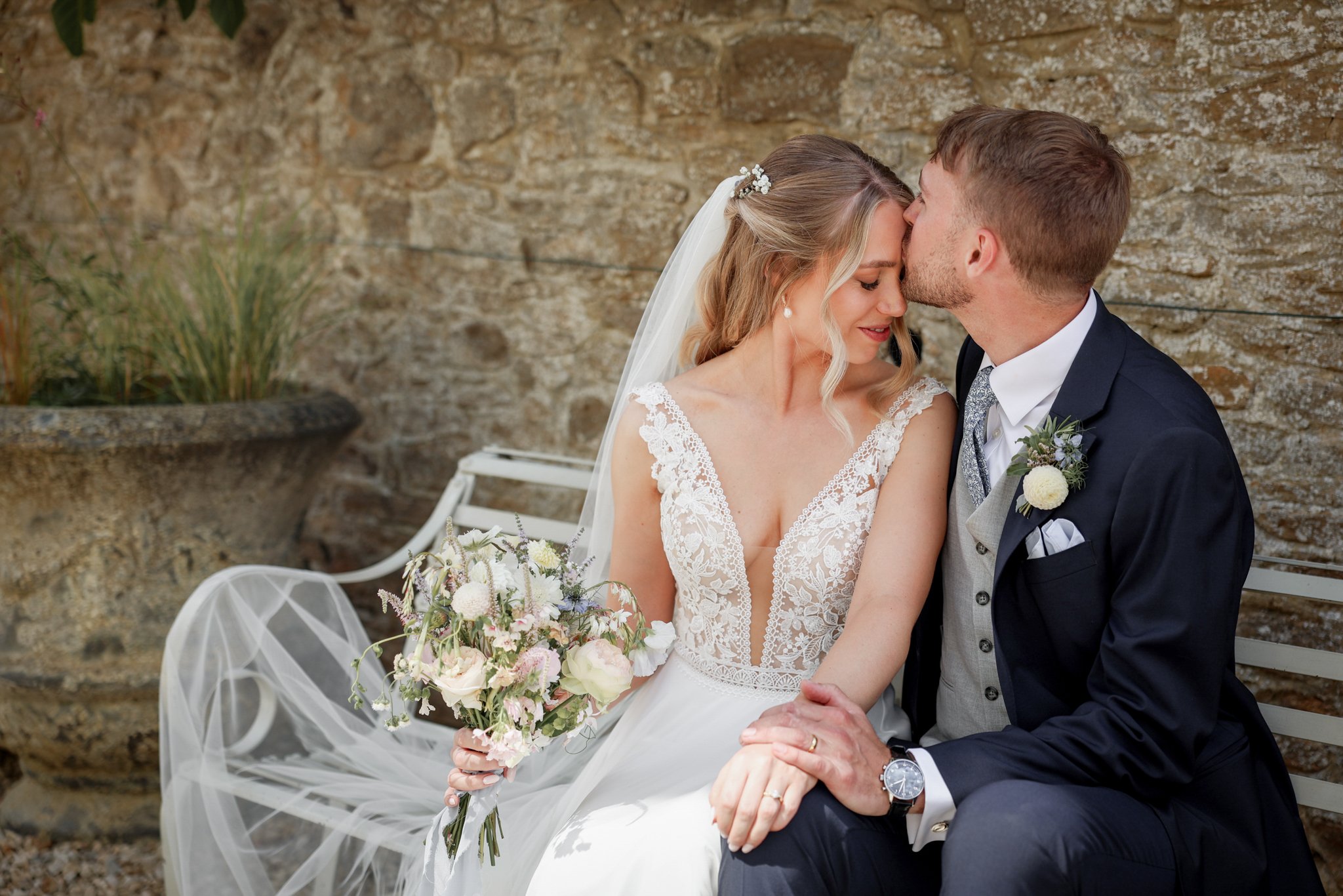 A bride and groom sitting closely together, about to kiss, against a stone wall. The bride is holding a bouquet of pink and white flowers, and they are sitting on a white bench with sheer white fabric draped over it.