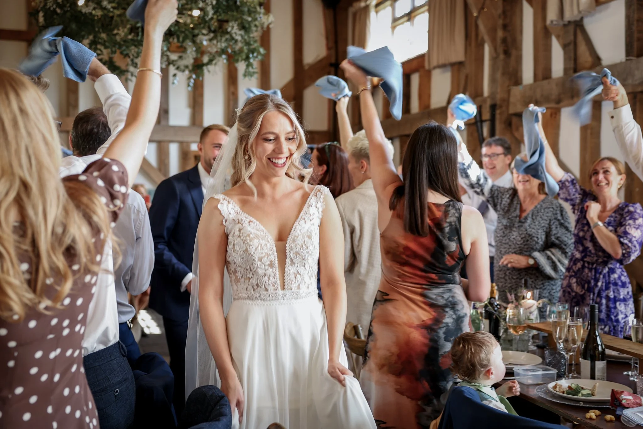A bride in a white wedding dress smiling with guests around her at a wedding reception in a rustic barn setting, some holding blue napkins in the air.